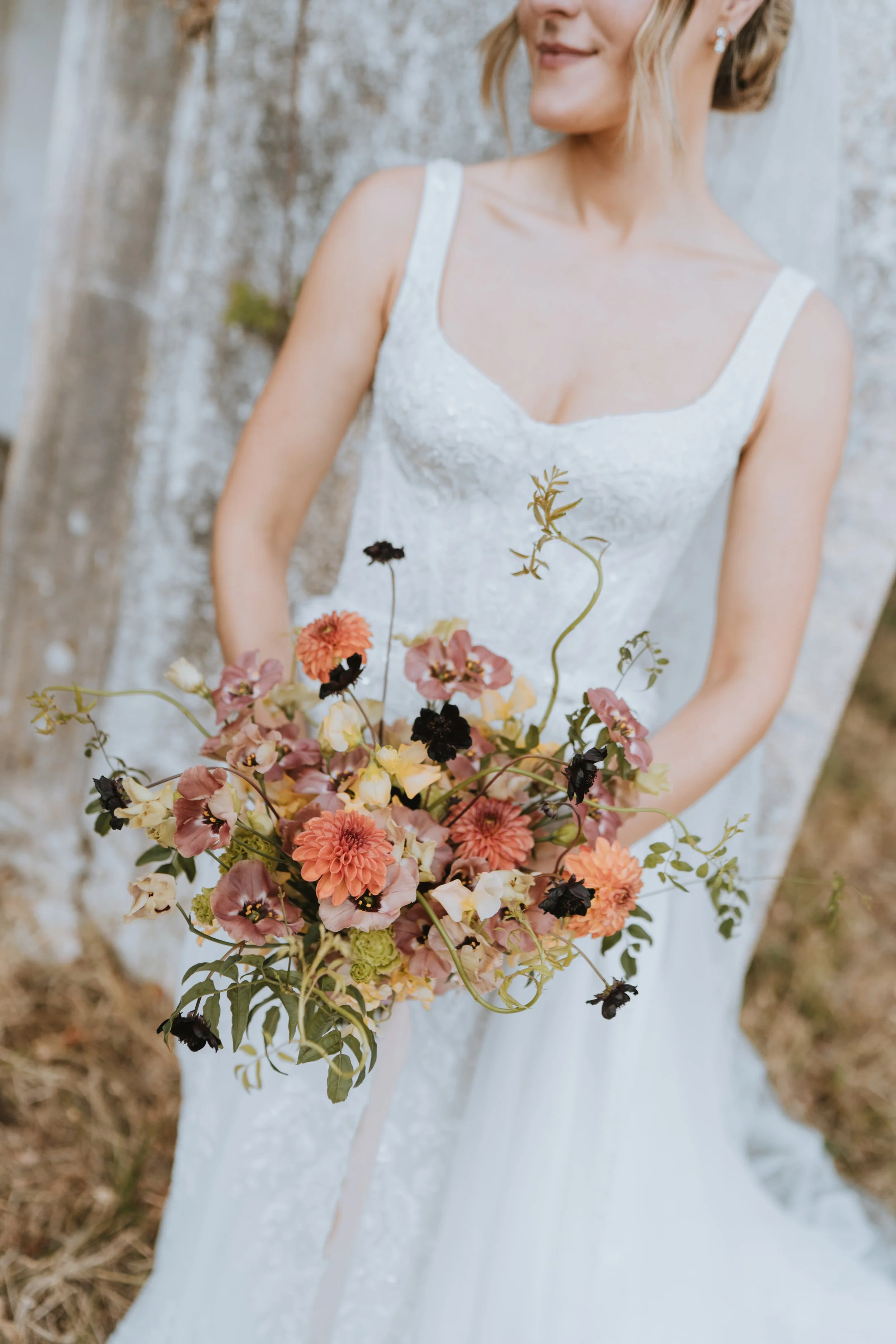A woman in a white wedding dress holds a bouquet of pink, peach, and black flowers, standing outdoors next to a tree.