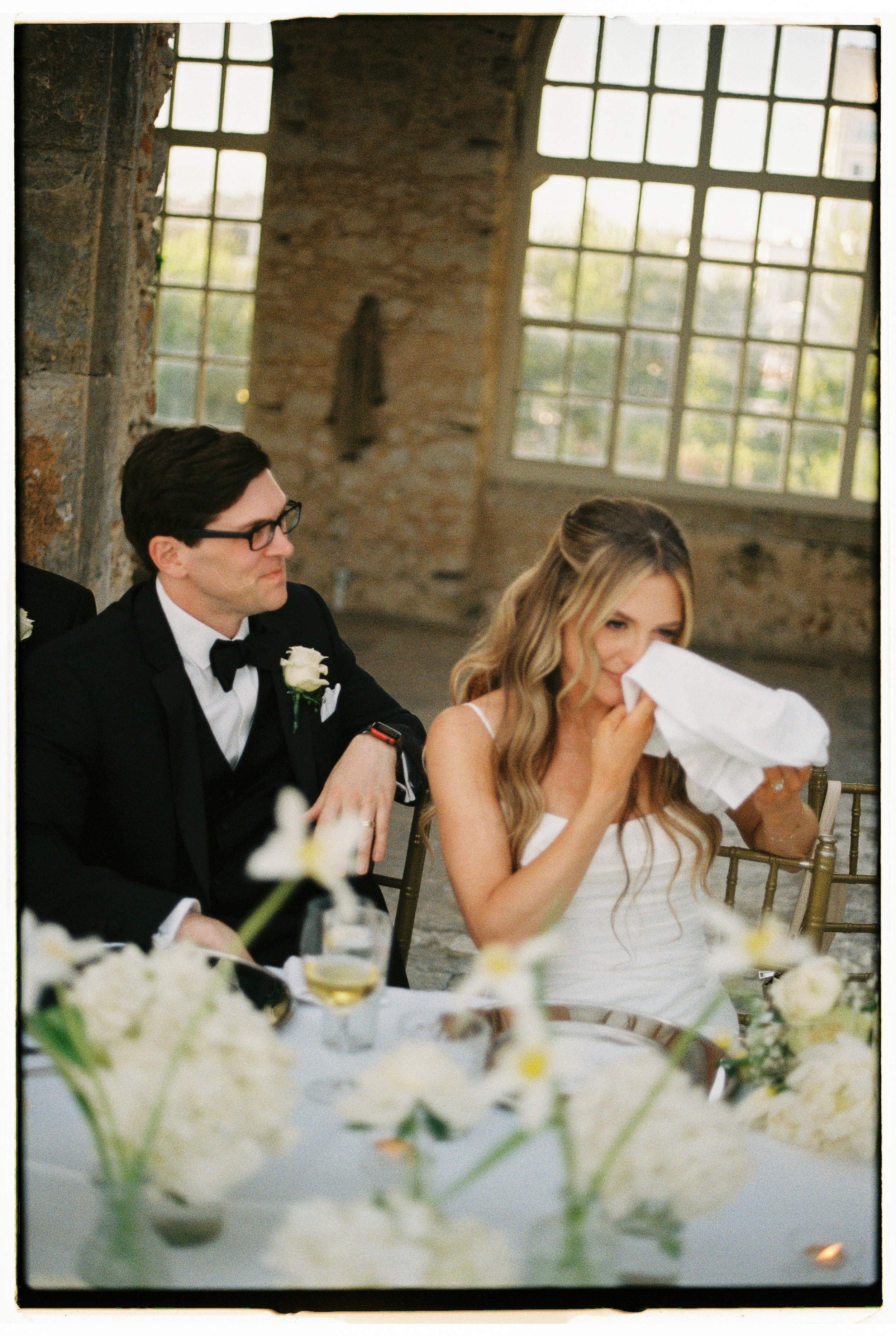 A bride in a white dress crying into a white napkin while sitting next to a groom in a black tuxedo at a wedding reception.
