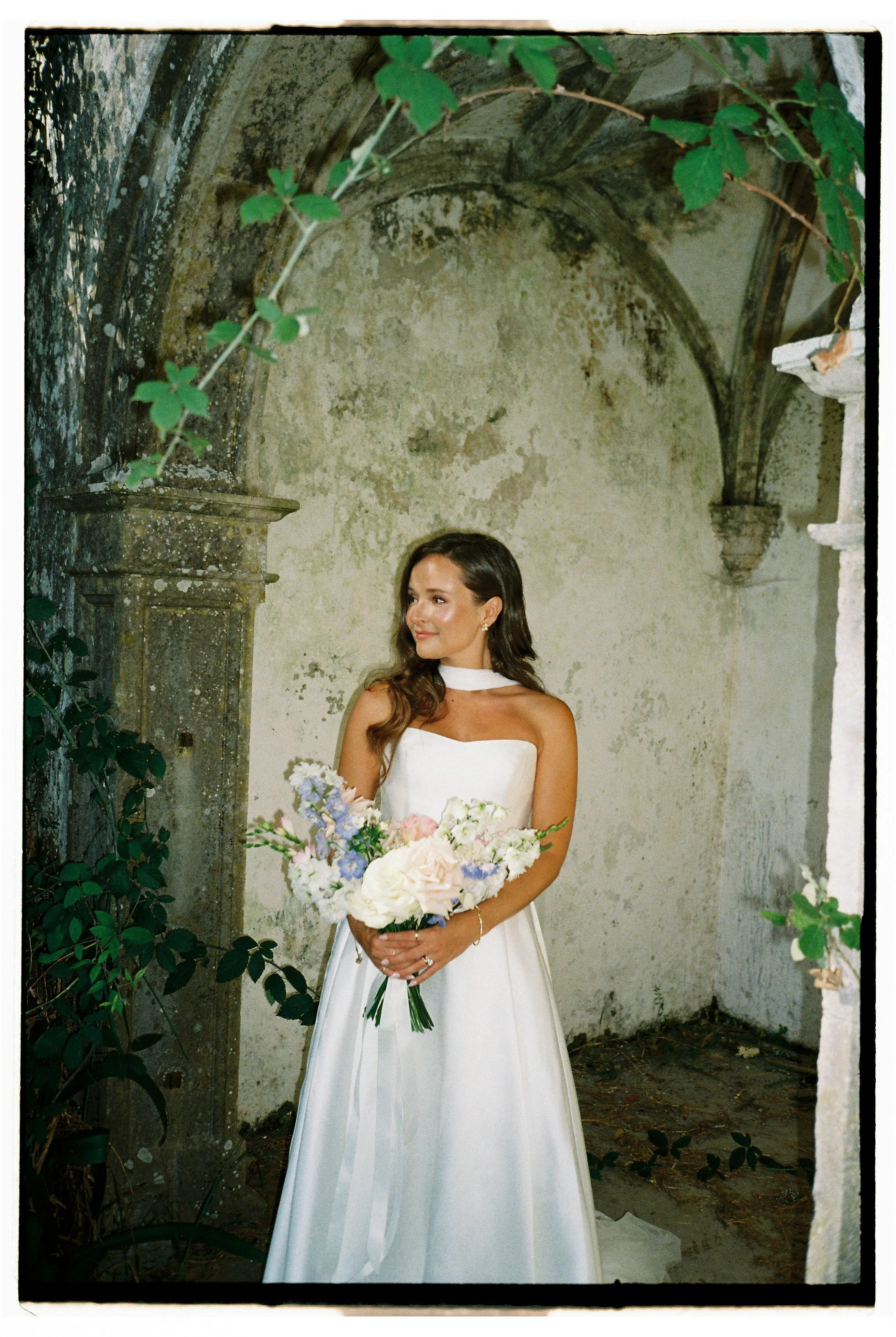 A woman in a white wedding dress holding a bouquet of flowers, standing in an old, rustic, stone-walled room with greenery and arches.