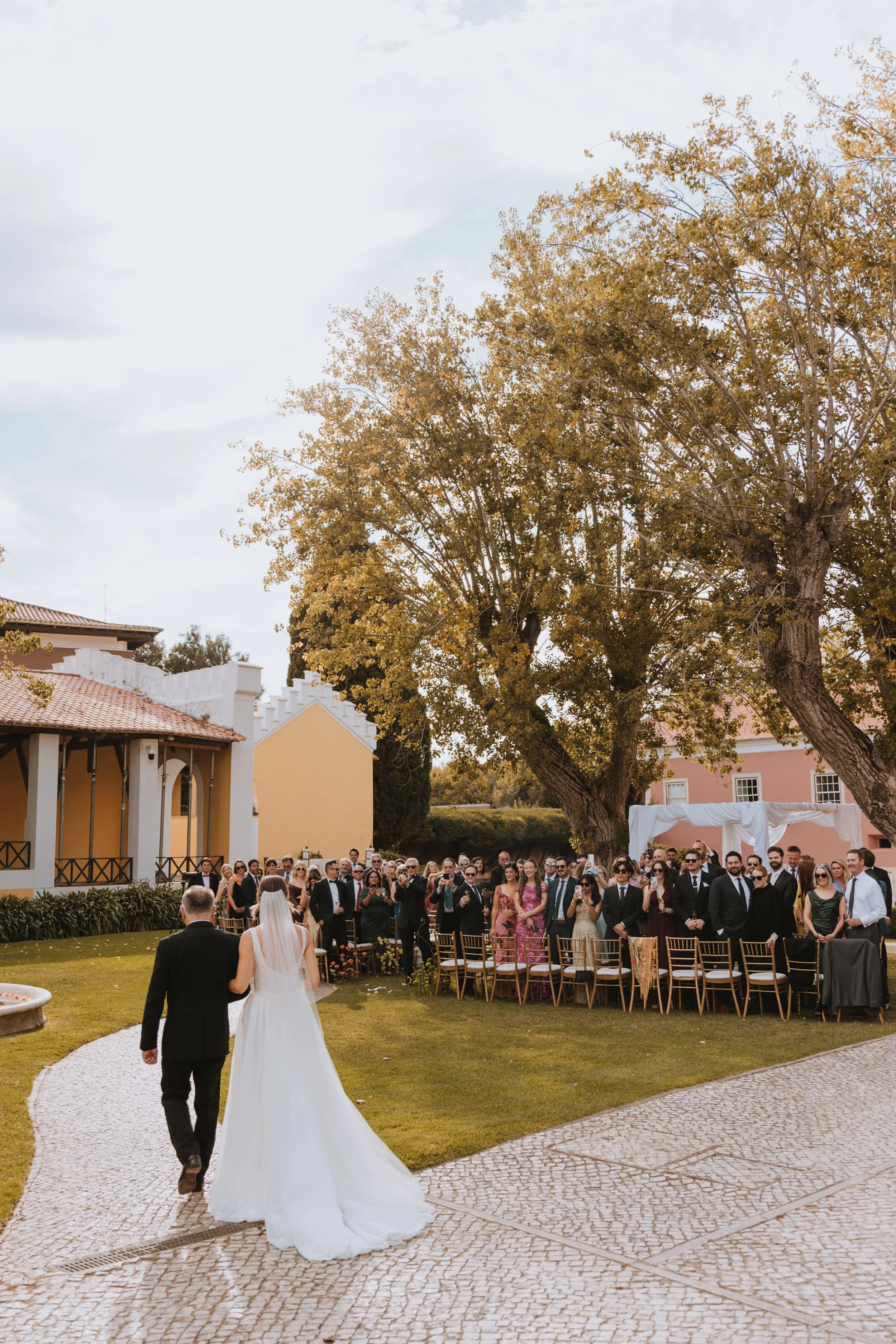 Bride and groom walking to the ceremony area with guests seated in an outdoor garden setting under large trees