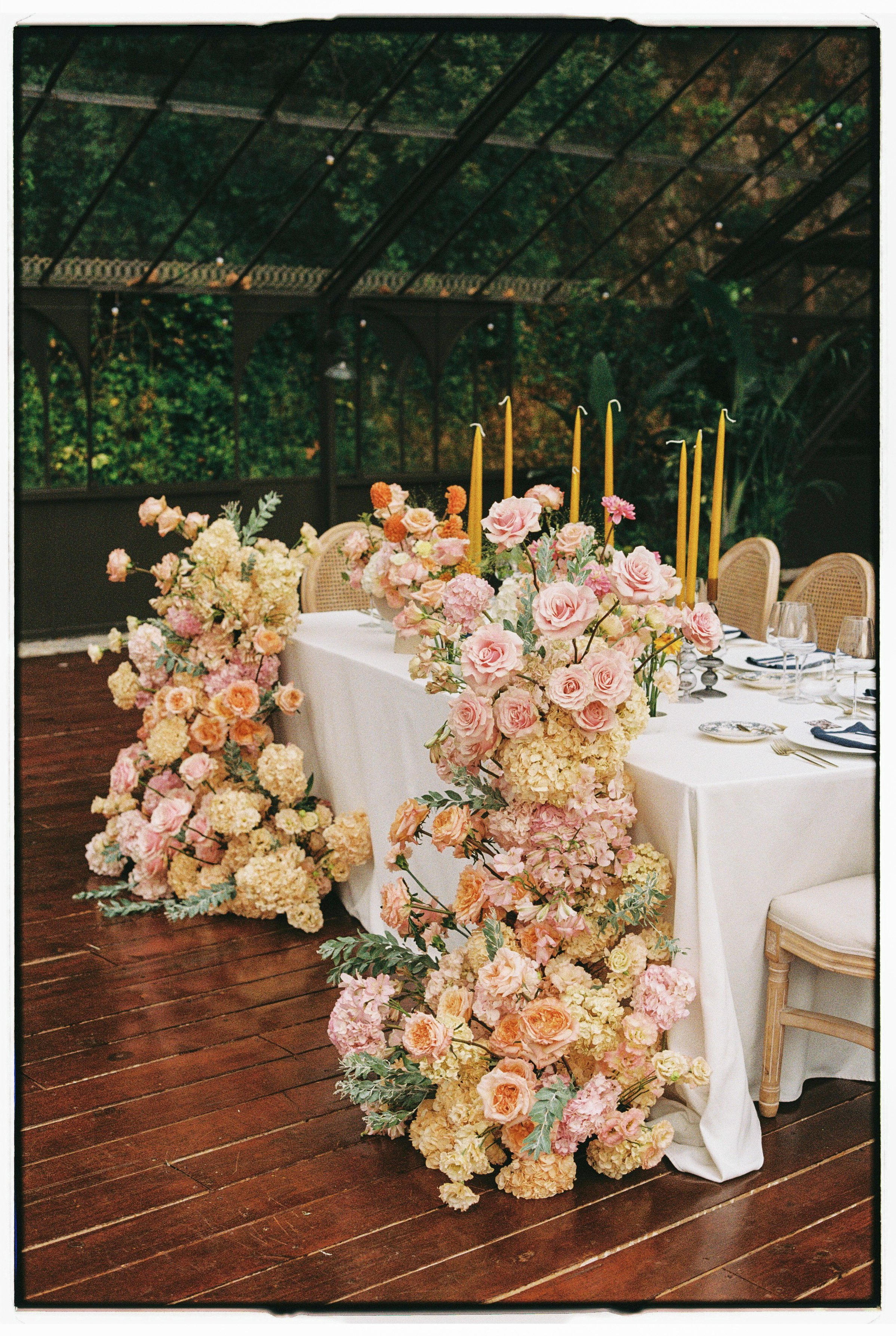 Elegant dining table decorated with pink and peach flowers, tall yellow candles, and formal place settings, set on a wooden floor with a garden or greenhouse background.