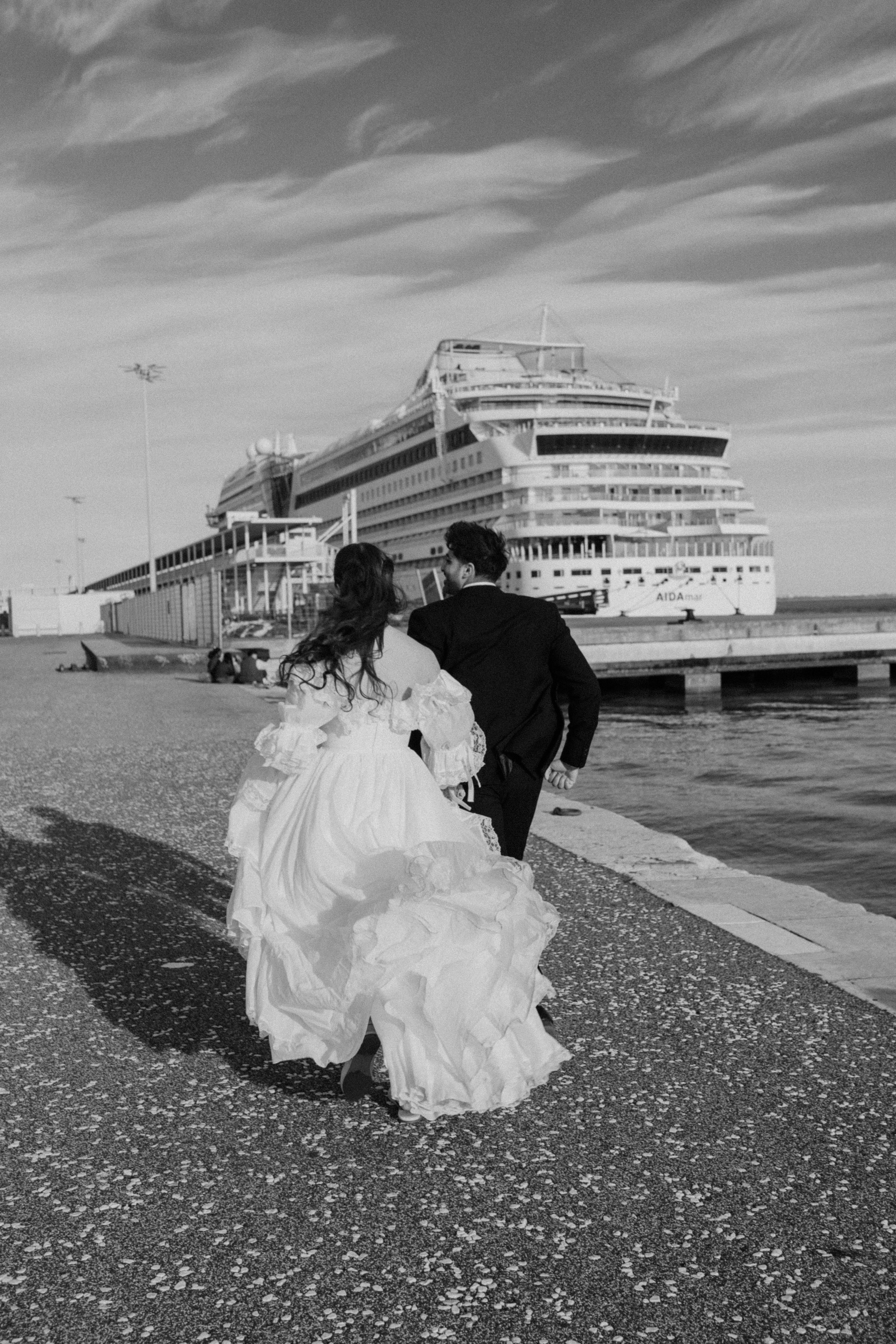 A couple dressed in wedding attire running along a pier with a large cruise ship docked behind them.
