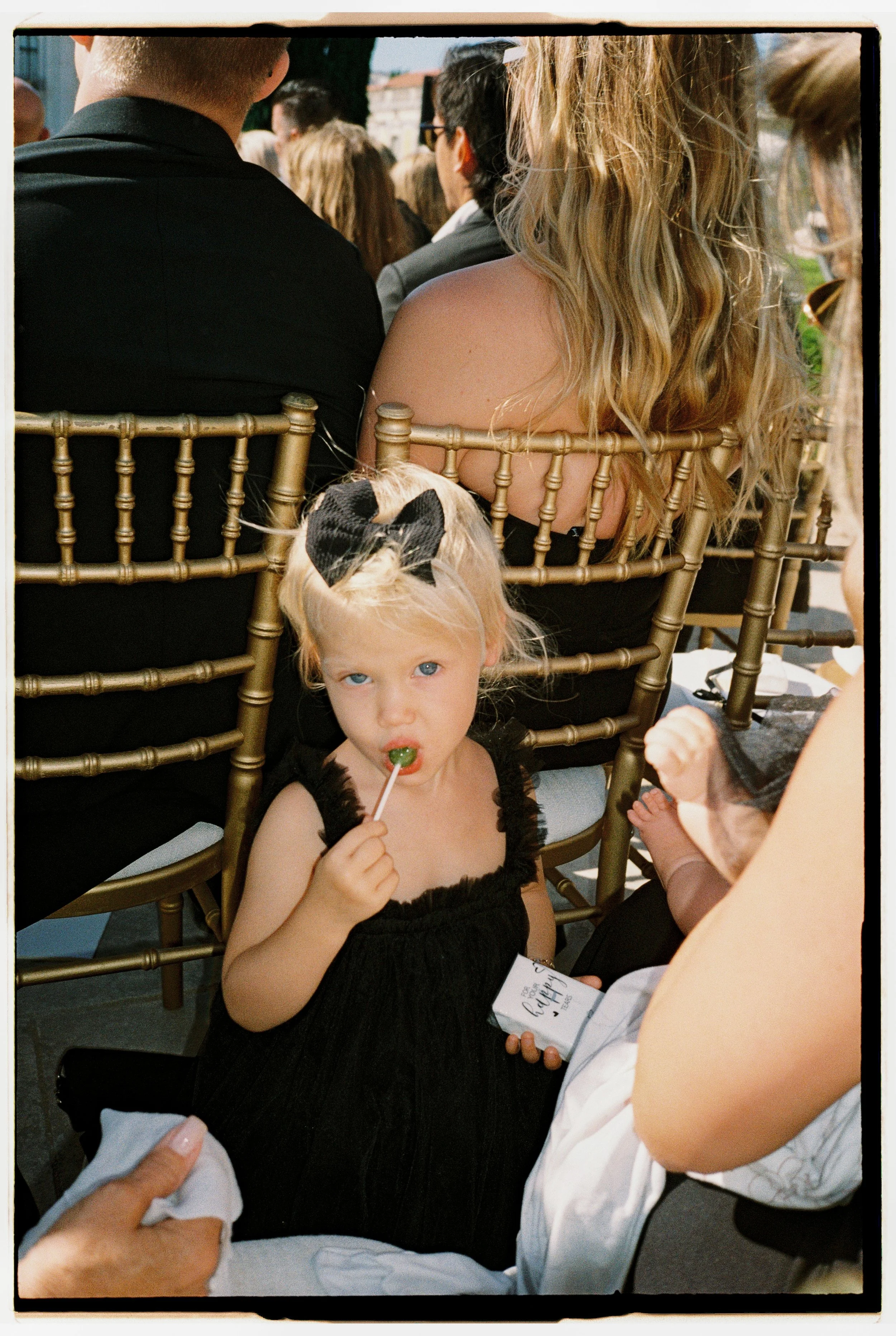 A young girl with blonde hair, blue eyes, and wearing a black dress with lace trim and a black bow in her hair, sitting among adults at an outdoor event. She is holding a green lollipop and looking directly at the camera.