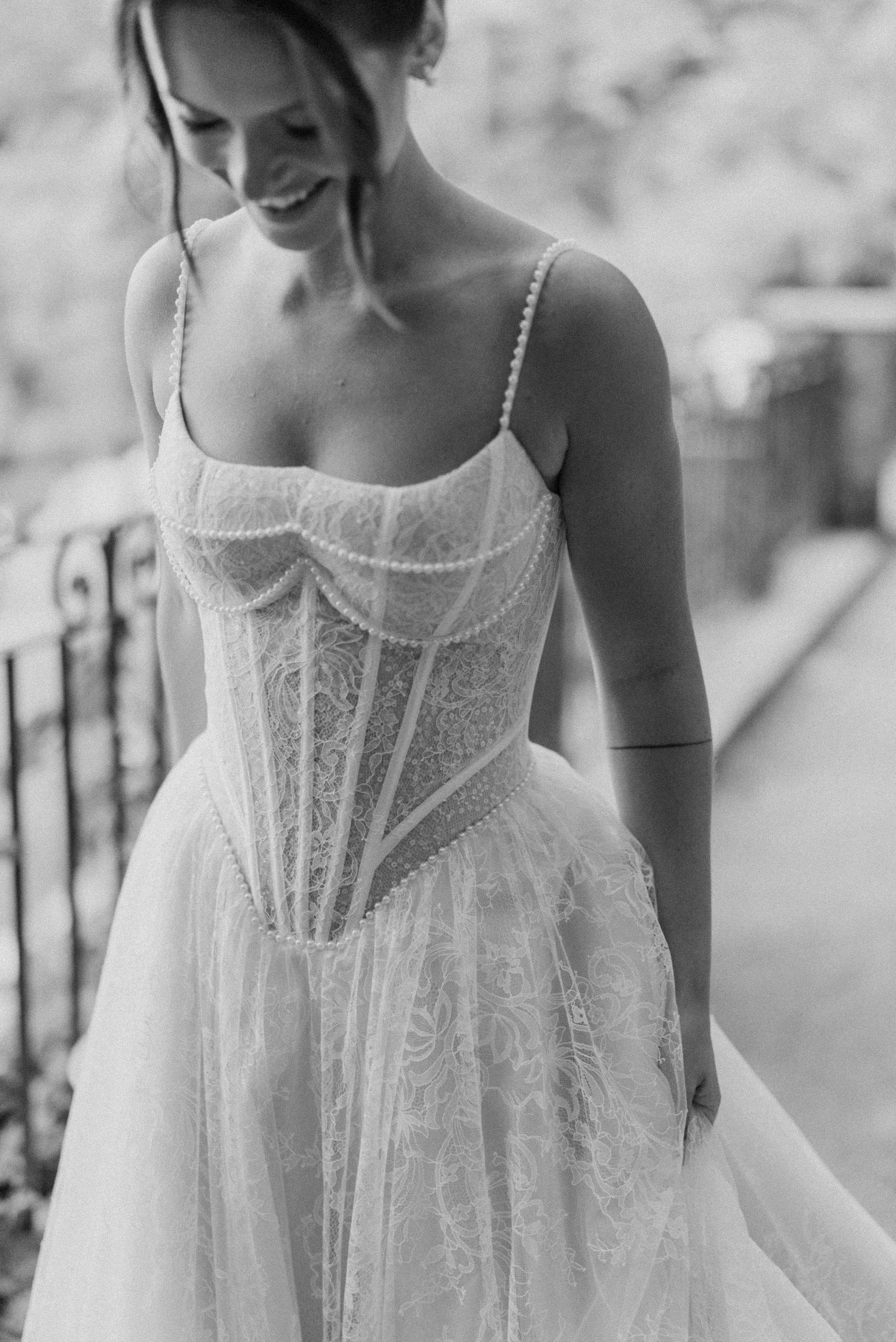 Black and white photo of a woman in a lace dress, smiling and looking down, standing outdoors near a railing.