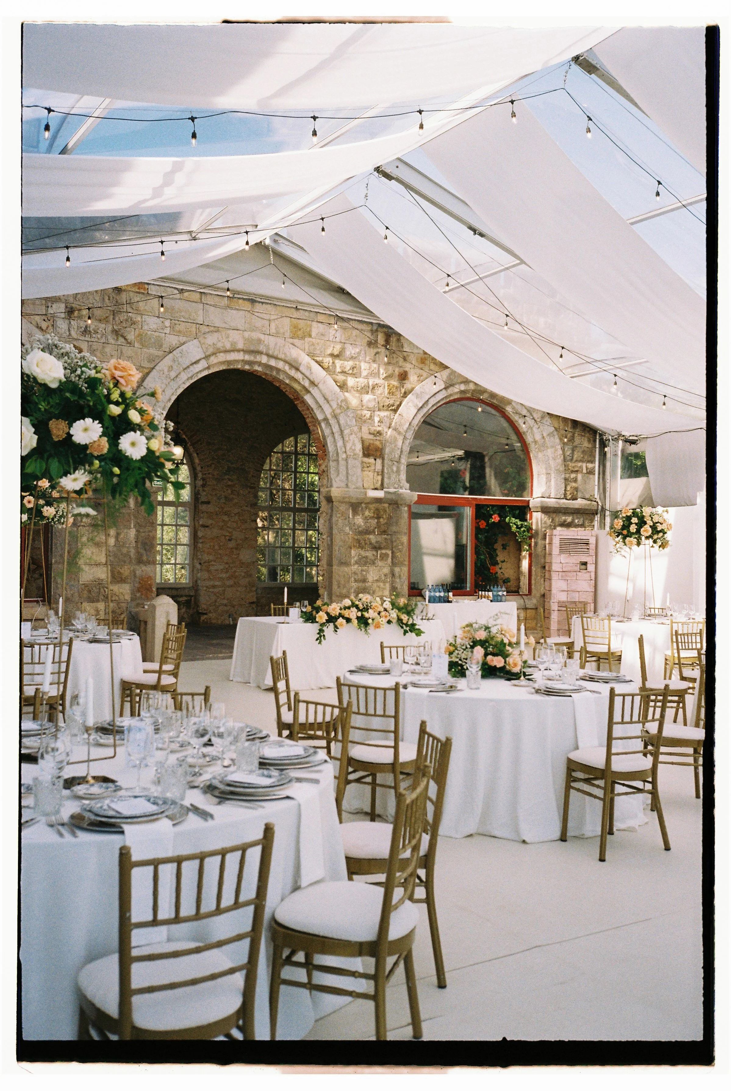 Decorated event space with round tables covered in white tablecloths, floral centerpieces, gold chairs, and a brick wall with arched windows, under white draped fabric and string lights.