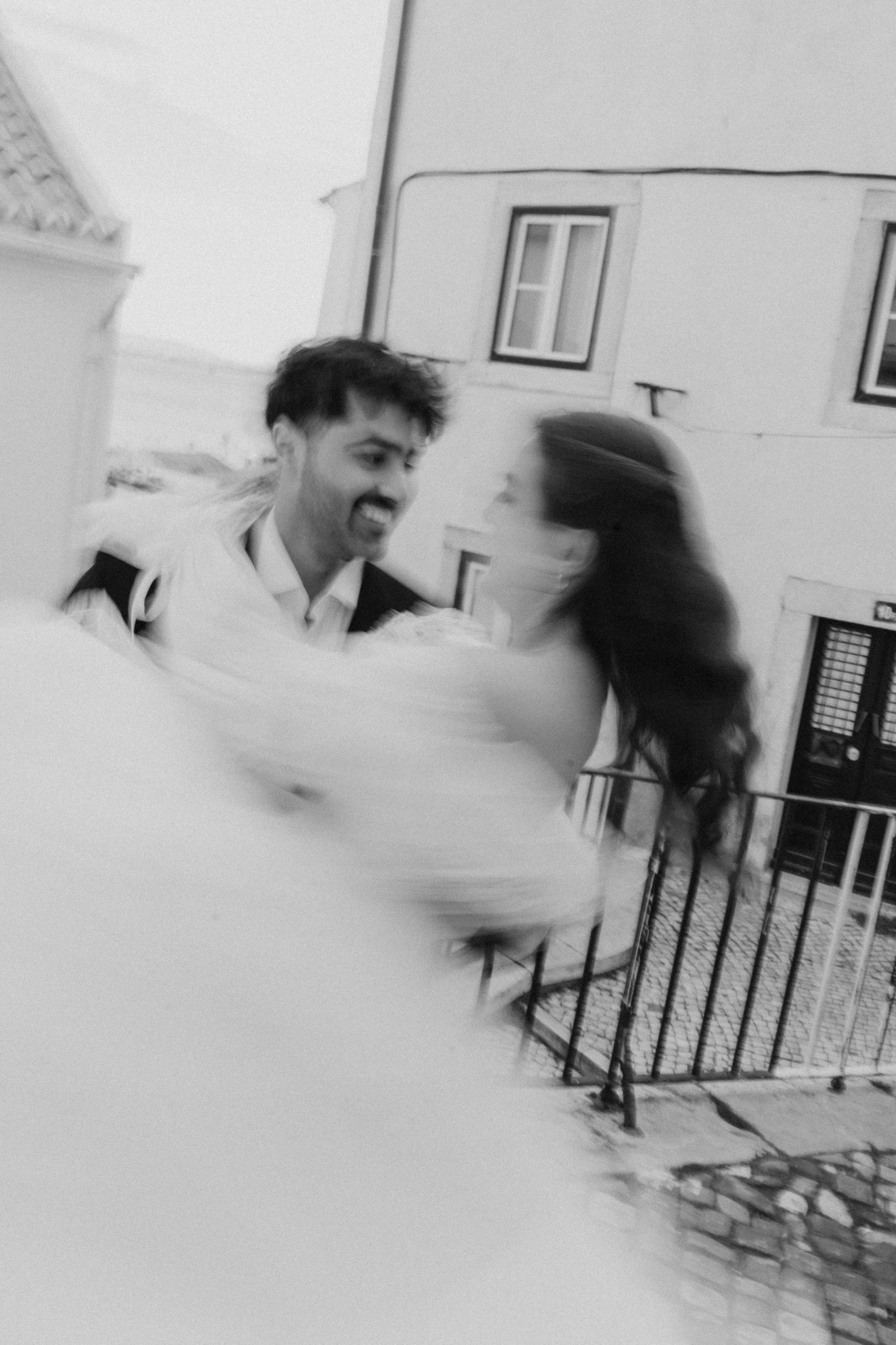 A black and white photo of a young man and woman embracing outdoors, smiling at each other with blurred motion around them.