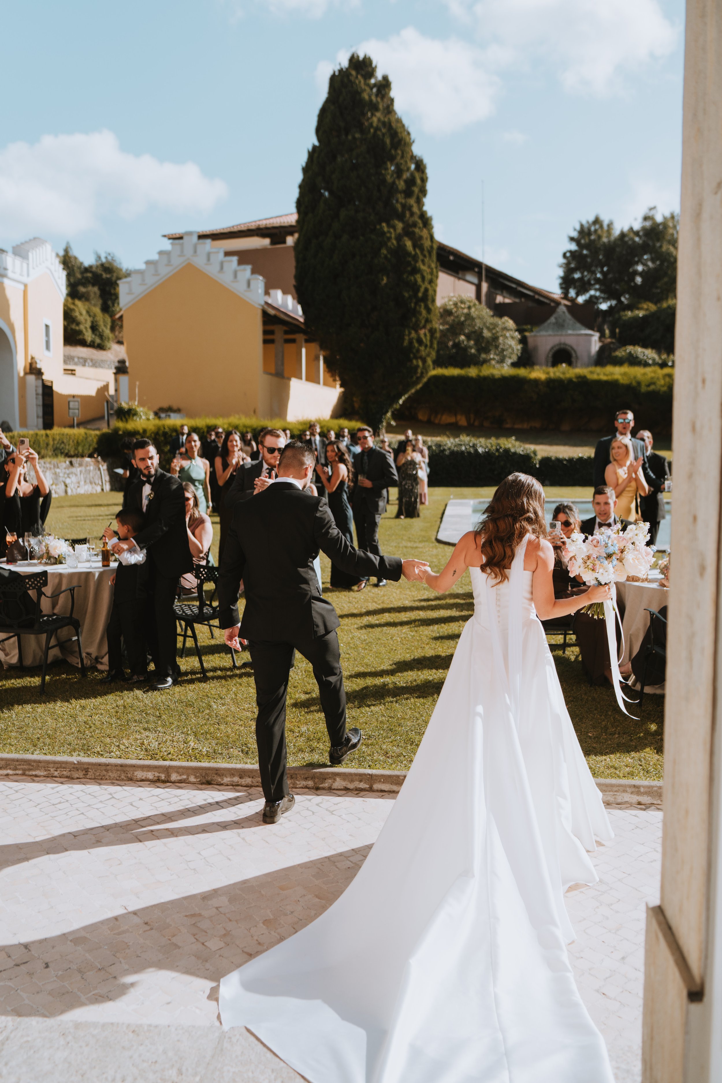 Bride and groom holding hands at their outdoor wedding reception, with guests seated and standing around in a lush garden setting.