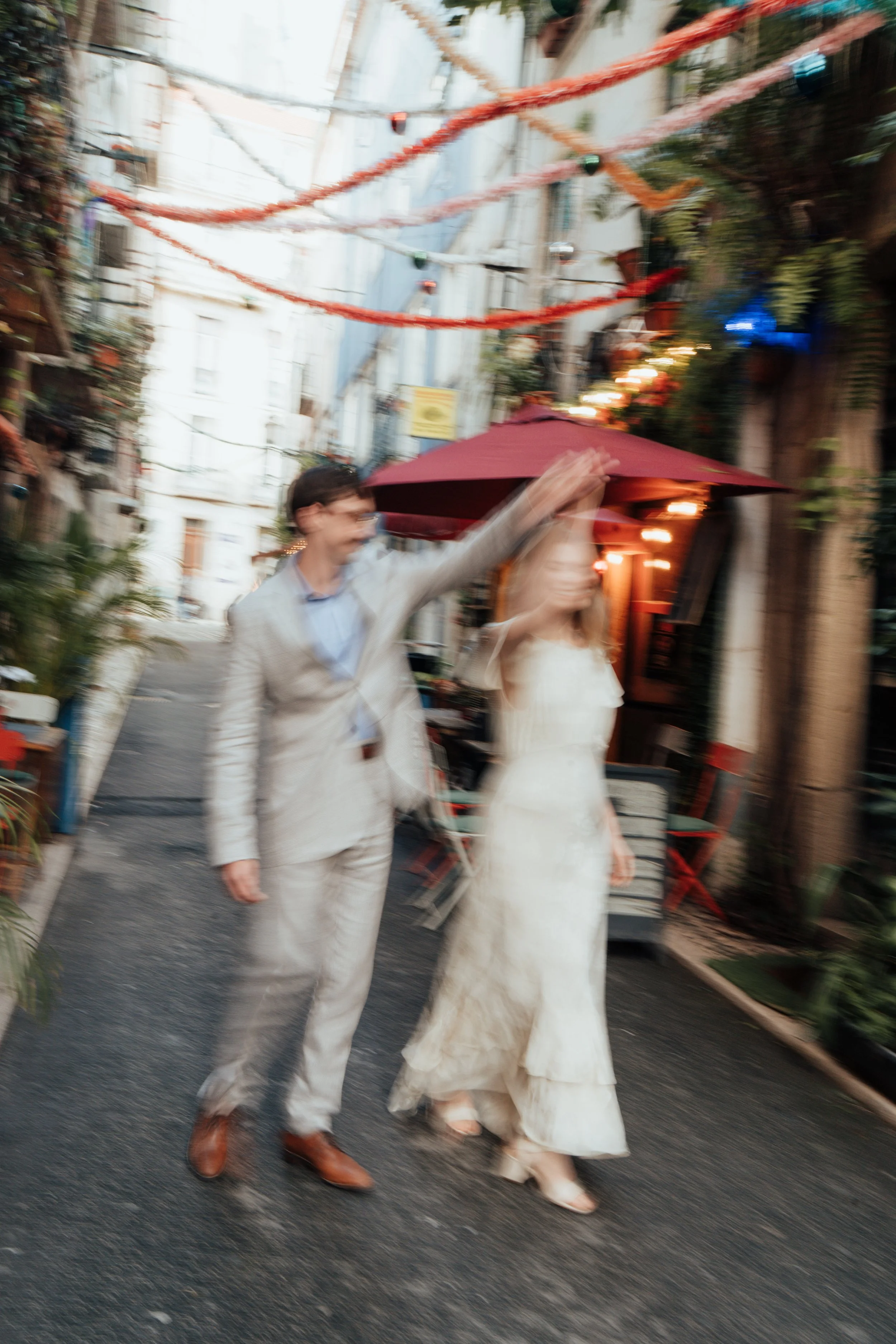 Blurry photo of a man in a light-colored suit with brown shoes and a woman in a white dress and heels, walking down a street decorated with hanging lights and plants, near a building with outdoor seating and a large red umbrella.
