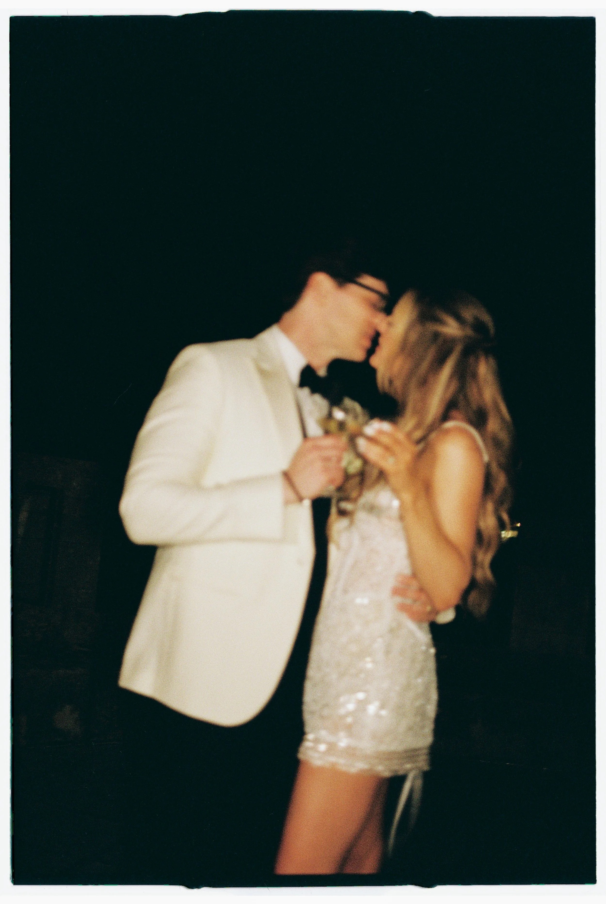 A romantic moment between a man in a white tuxedo and a woman in a lace dress, standing close with foreheads nearly touching, at a dark background event.