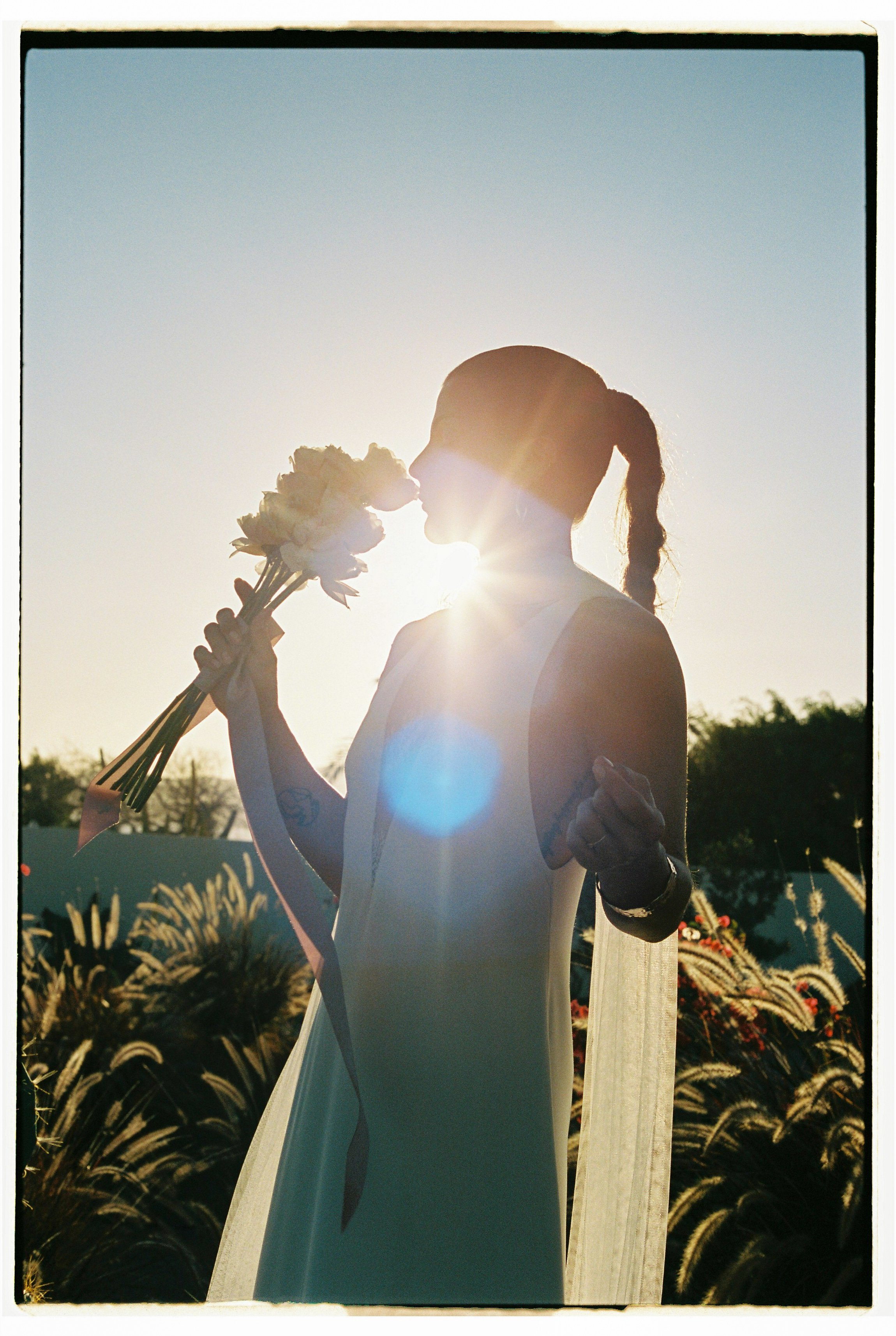 Silhouette of a woman in a white dress holding a bouquet of flowers close to her face, with sunlight shining behind her, creating a halo effect. She is outdoors with trees and tall grasses in the background.