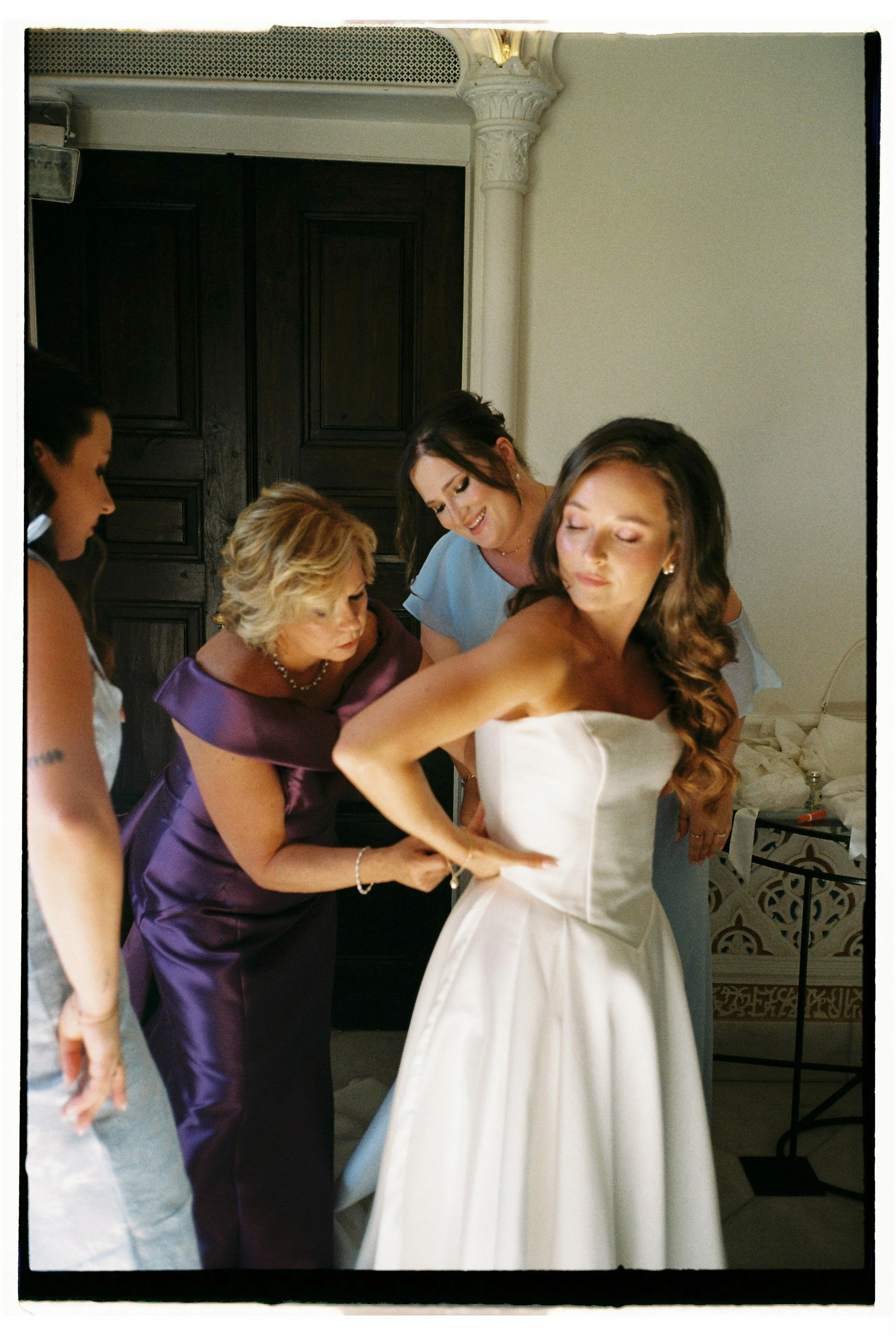 A bride in a strapless white wedding gown getting ready with four women around her, helping her dress or adjust her gown, in a room with a dark wooden door and ornate white columns.