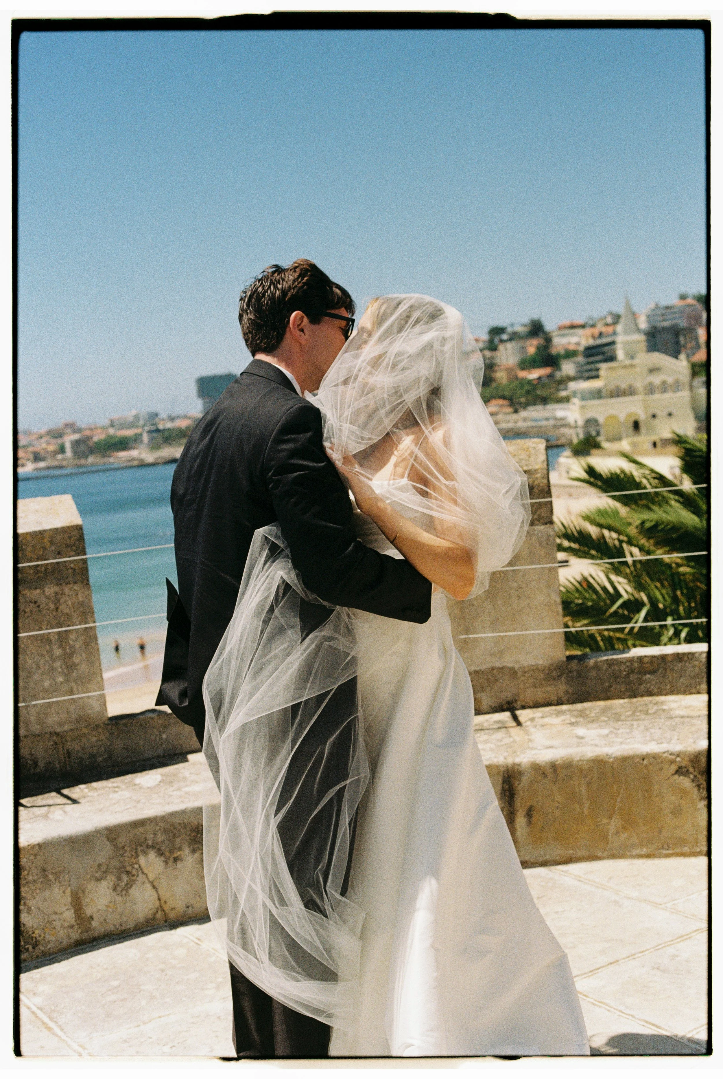 A couple on their wedding day, with the groom in a black tuxedo and the bride wearing a white wedding gown and veil, standing outdoors with a view of the water and city buildings in the background.