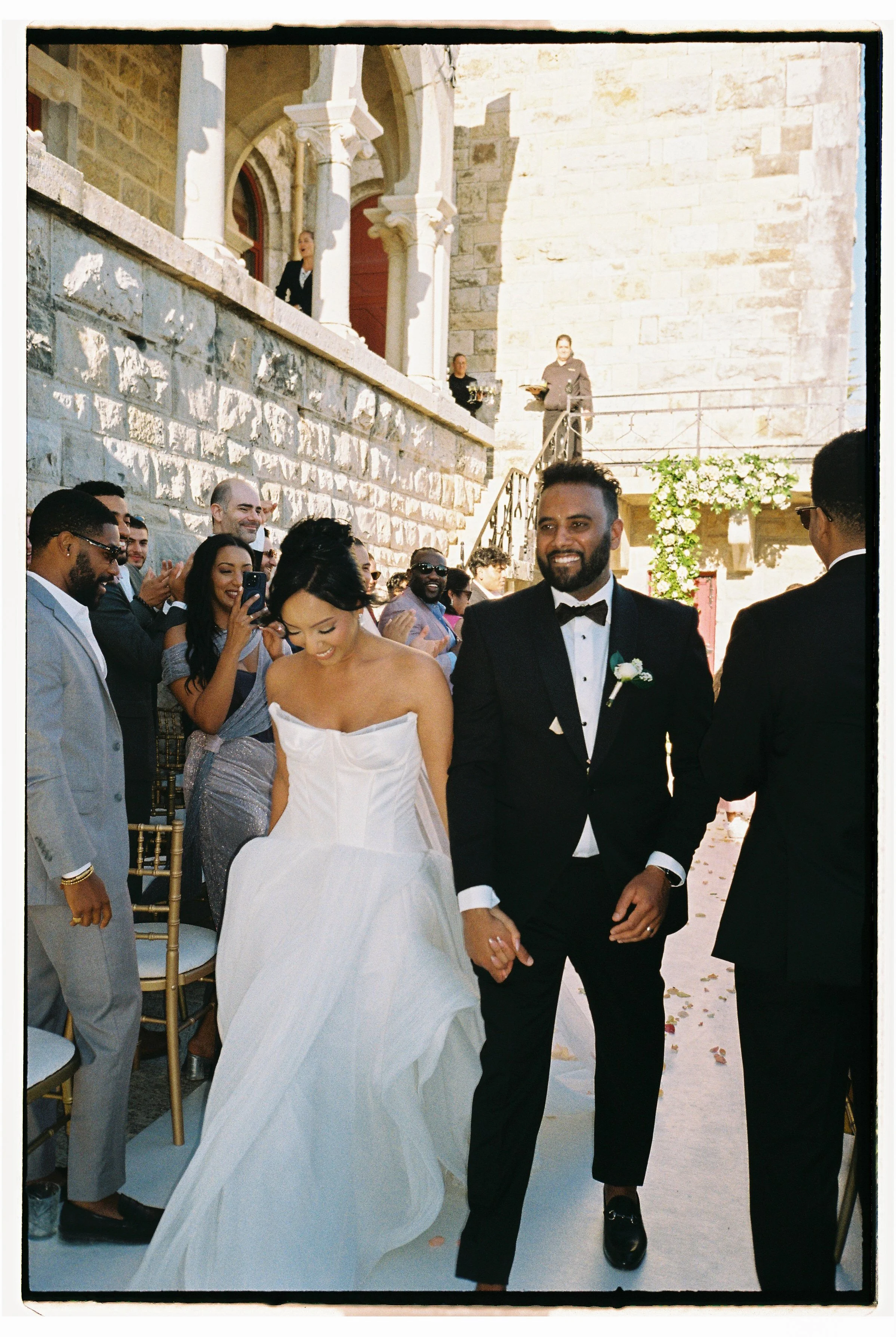 A newlywed couple walking down the aisle at their outdoor wedding ceremony, surrounded by smiling guests, with a stone building and floral decorations in the background.