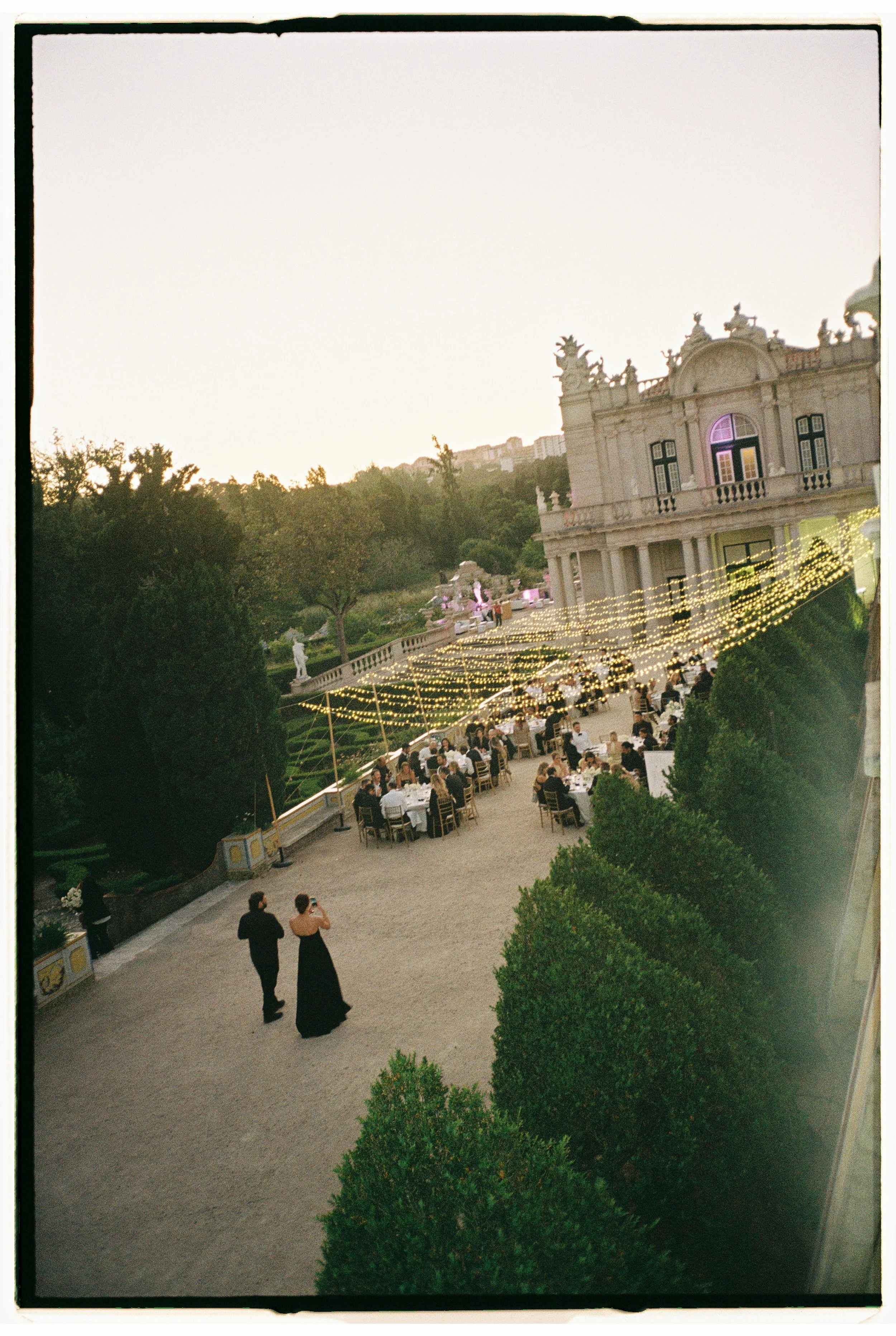 An outdoor wedding reception with tables of guests under string lights, set against a historic building with ornate architecture, greenery, and a couple in formal attire walking on a gravel path.
