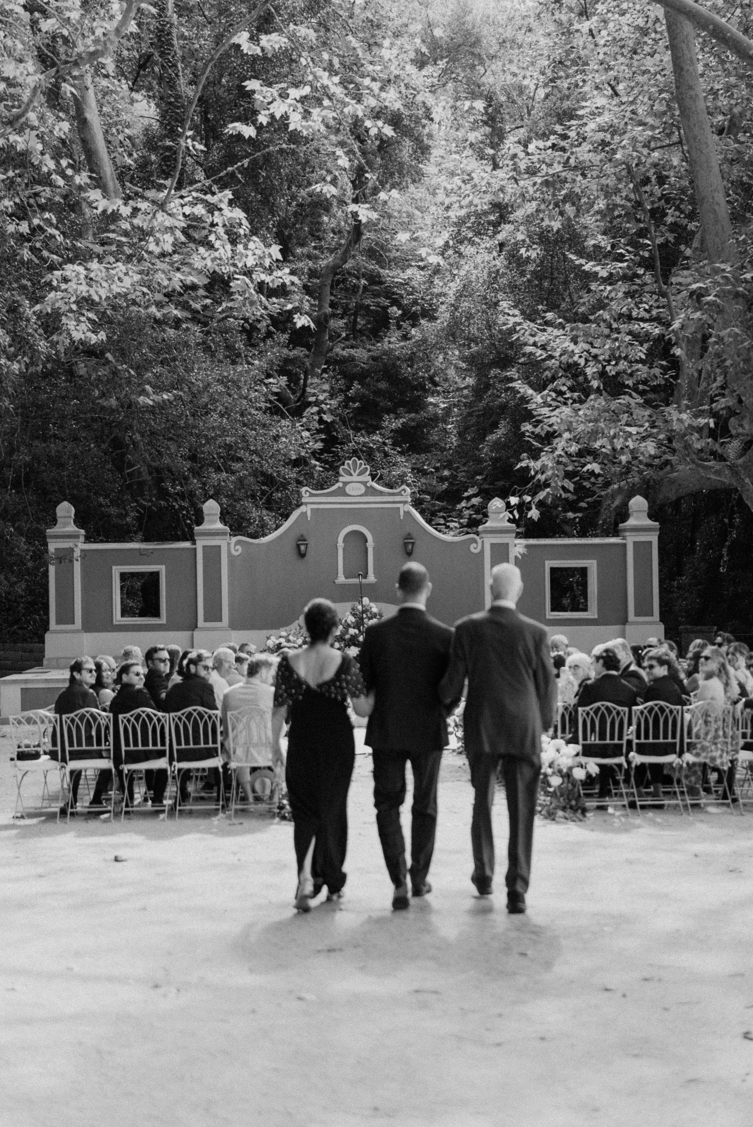 A couple walking down the aisle at an outdoor wedding ceremony held in a wooded area. The scene is in black and white.
