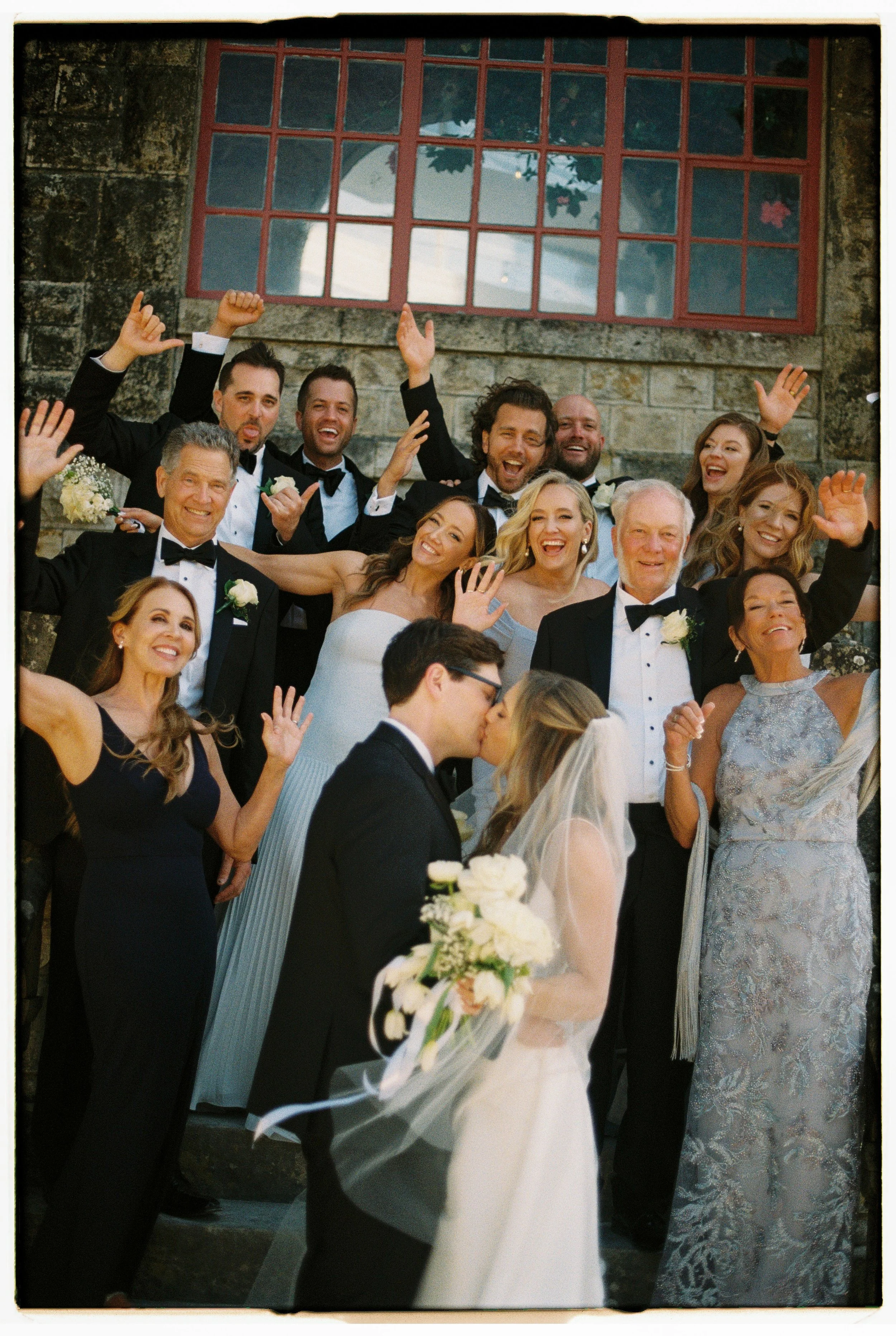 A wedding celebration with a bride and groom kissing in front of a large group of smiling guests, including children and adults in formal attire, outside a stone building with a large window, as guests cheer and wave.
