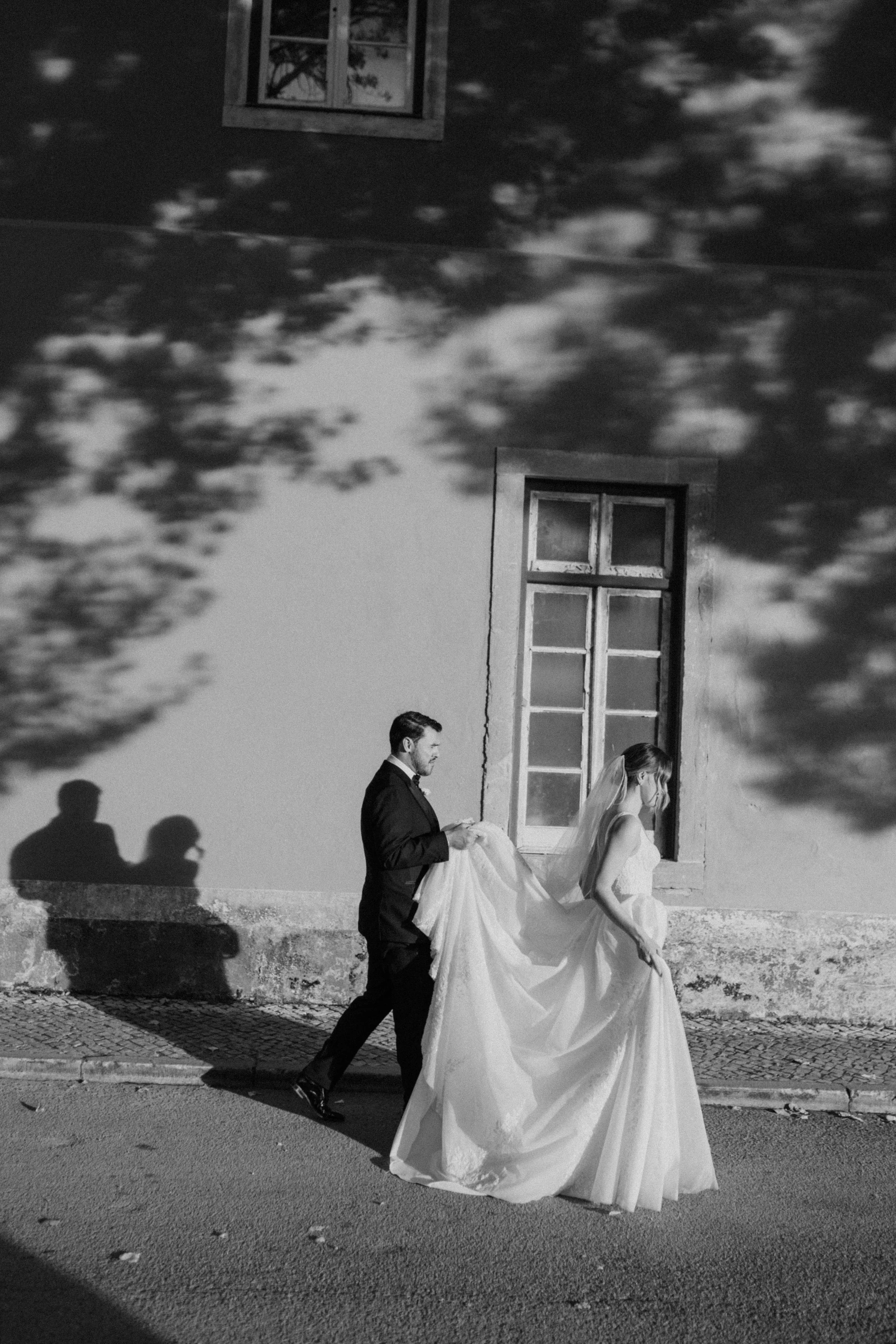 Black and white photo of a bride and groom walking past a building with a window. The groom is holding the bride's gown as they walk, with their shadows cast on the wall behind them.