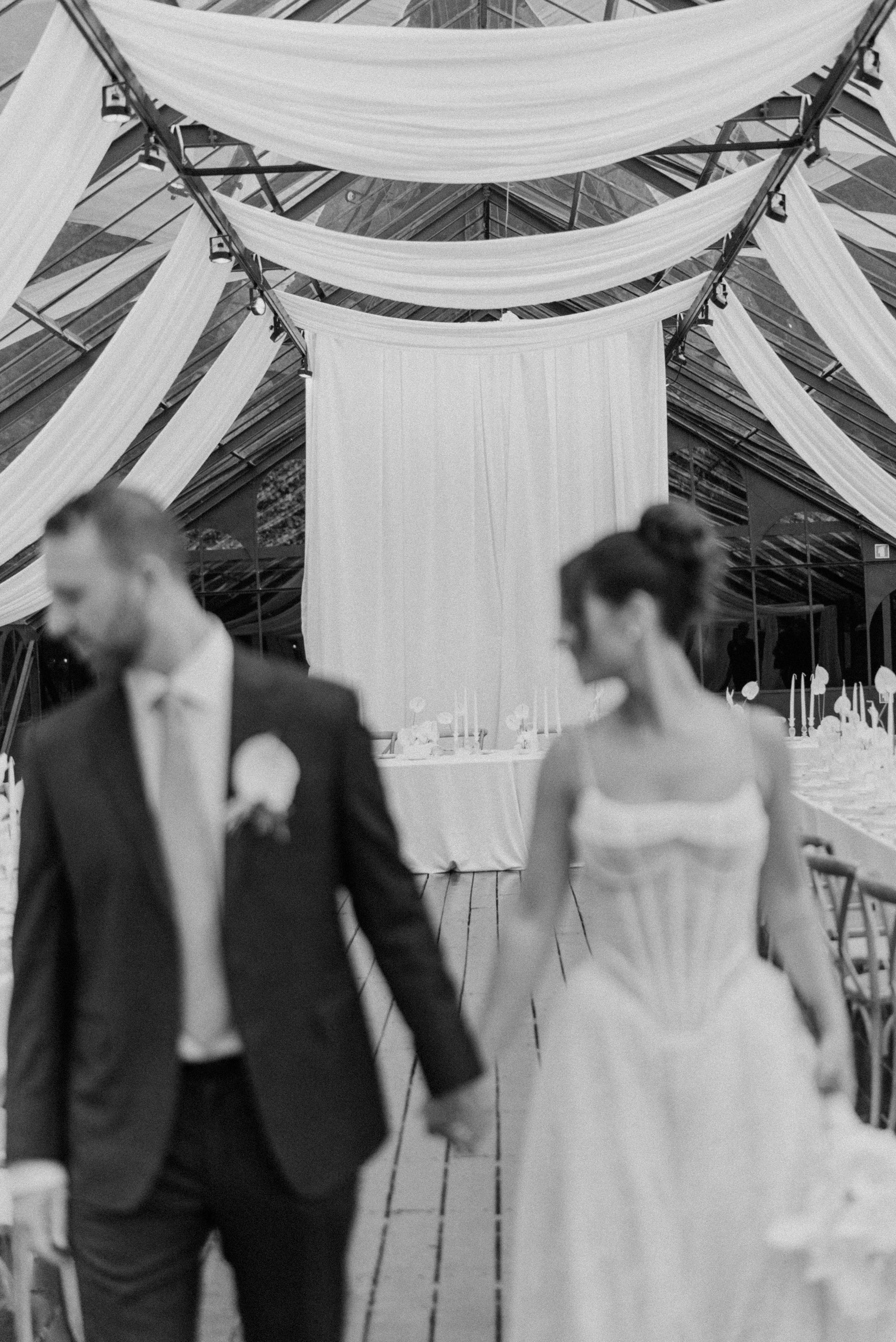 A black and white wedding reception with a bride and groom holding hands and walking together, decorated with draped fabric and candle-lit tables.
