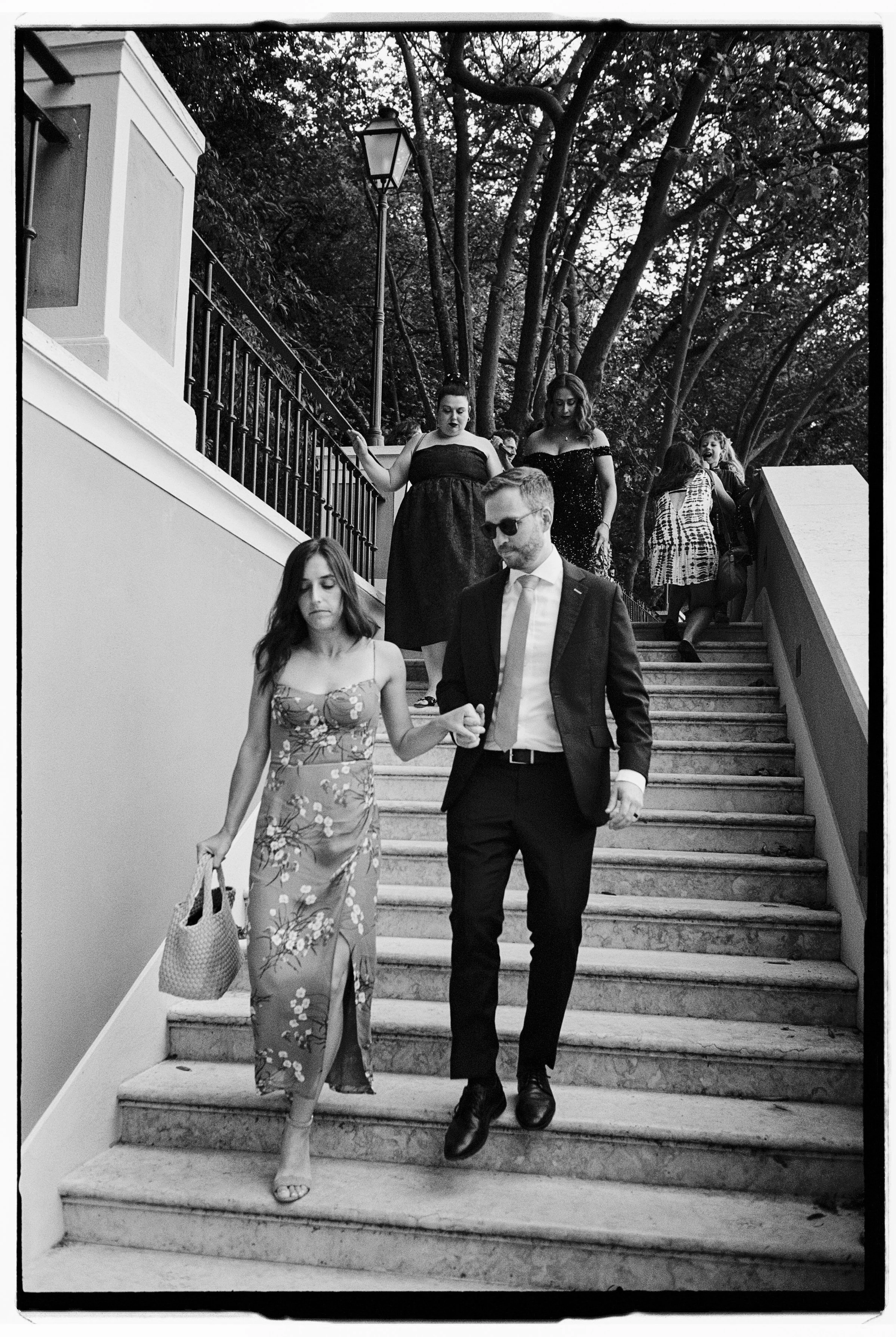 A group of people, dressed formally, descending outdoor staircase surrounded by trees and foliage, with a streetlamp visible above.