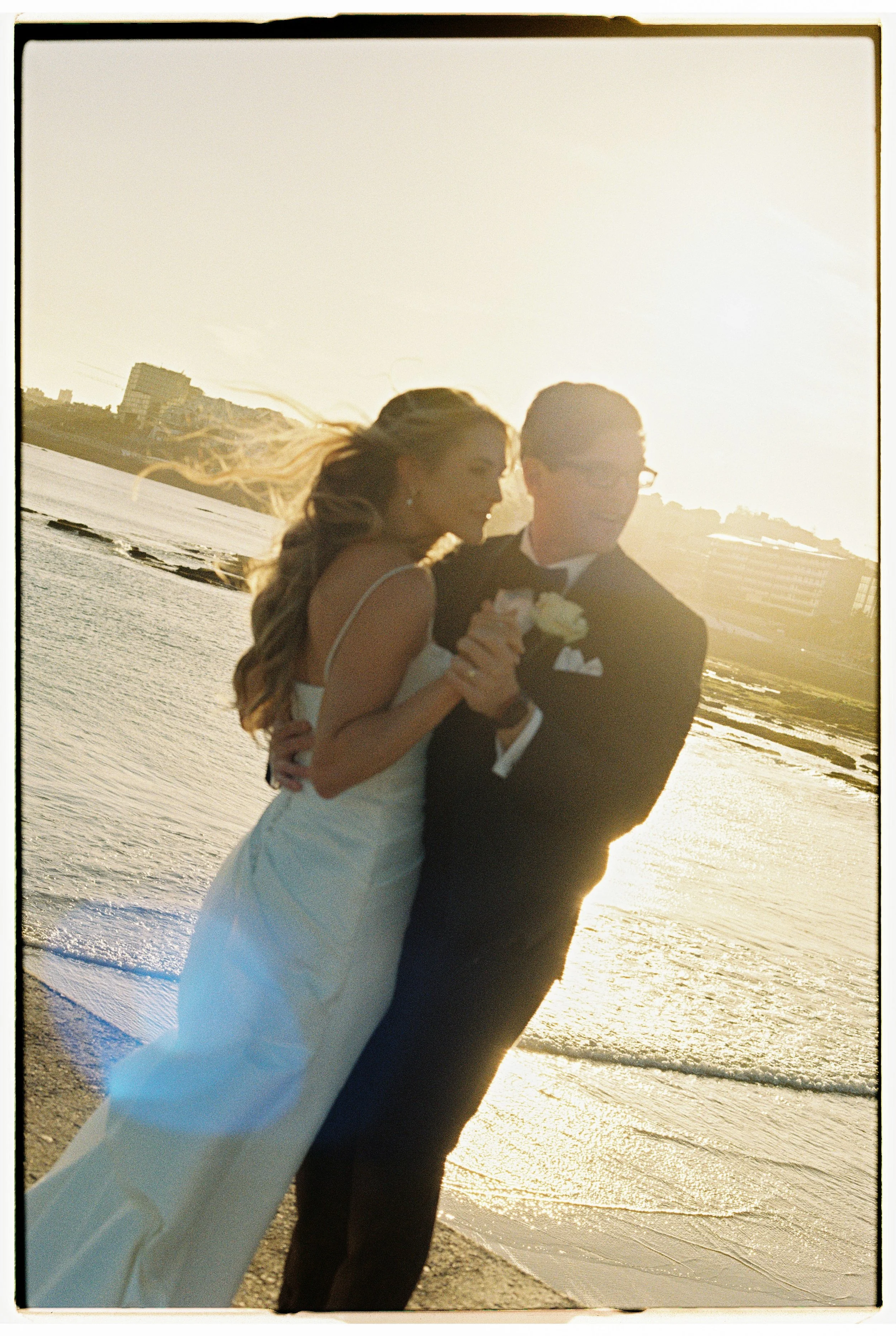 A couple in wedding attire standing on the beach at sunset, holding hands and smiling.