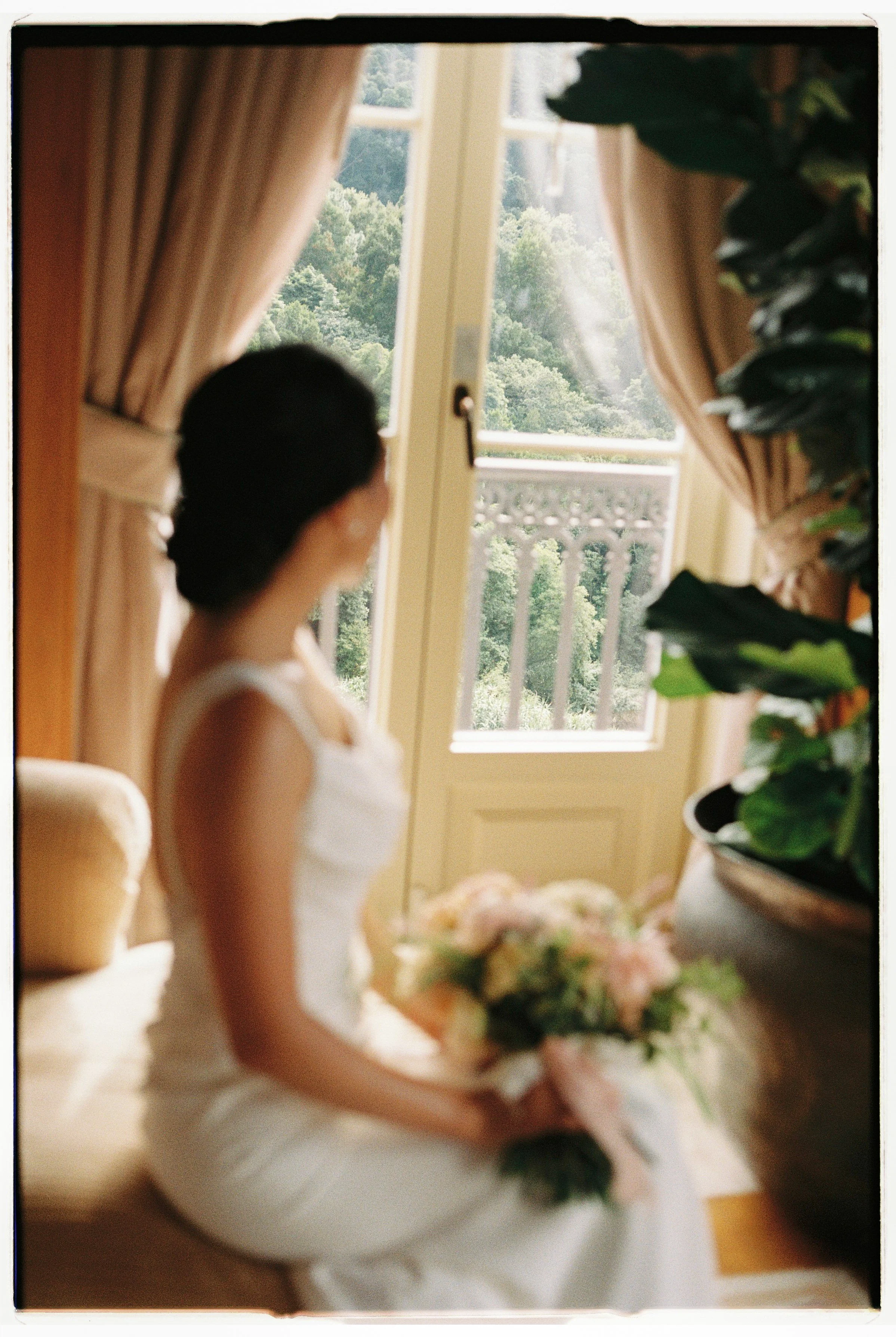 A woman in a white dress sitting near a window, holding a bouquet of flowers, with lush green trees visible outside.
