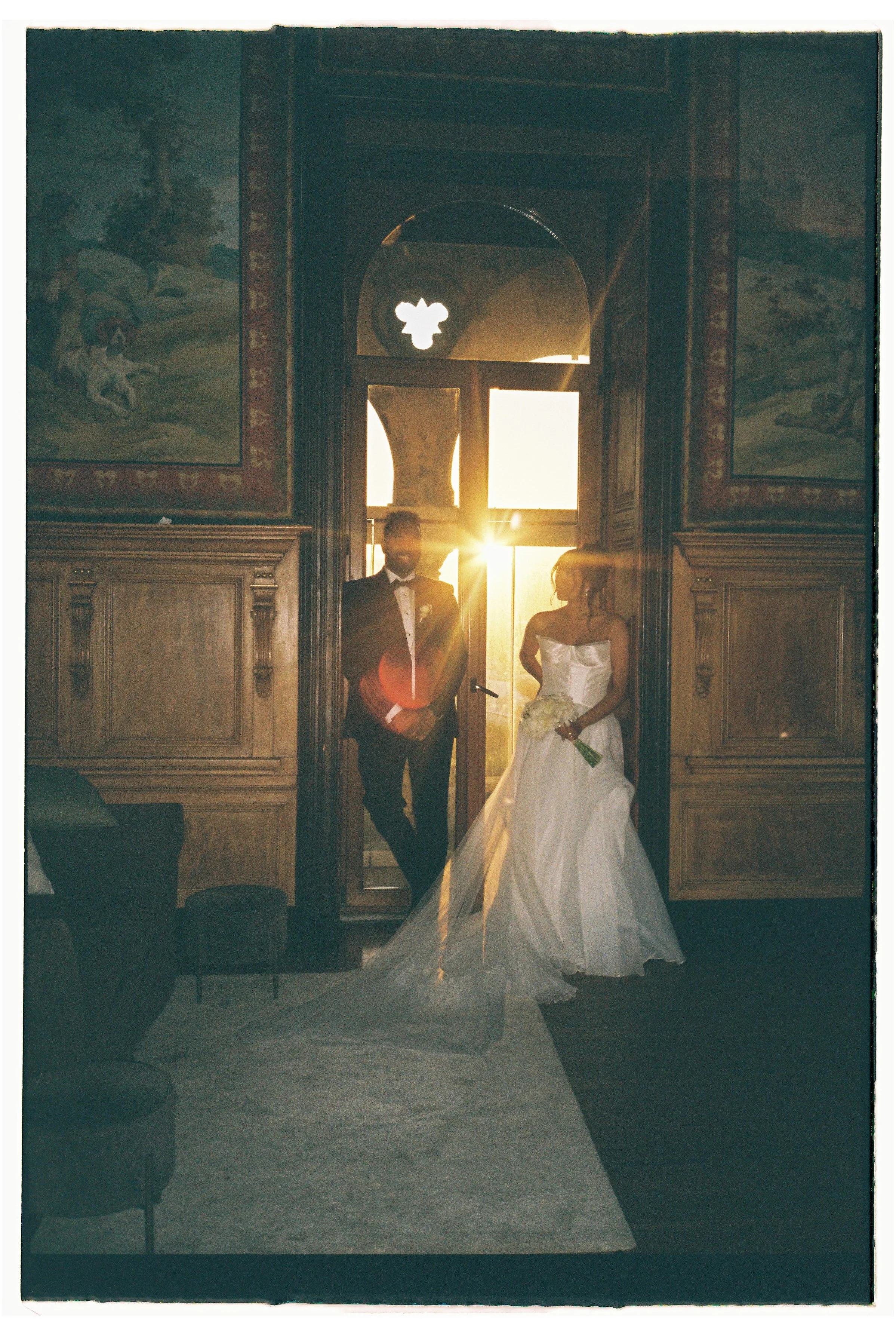 A bride and groom standing in a room with ornate wood paneling and murals, backlit by a bright setting sun through a large doorway, creating a silhouette effect.