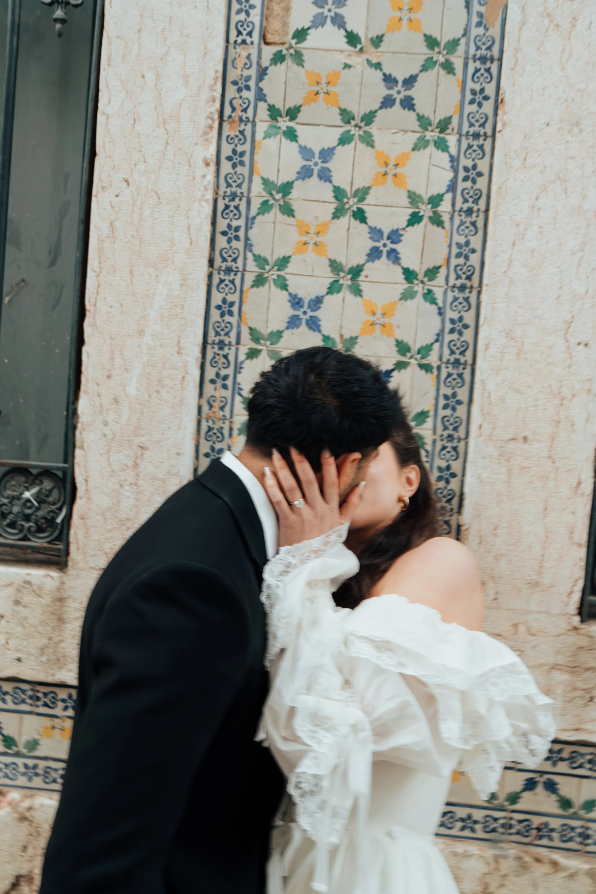 A couple sharing a kiss, with the woman's hand on the man's face, in front of a decorative tiled wall.