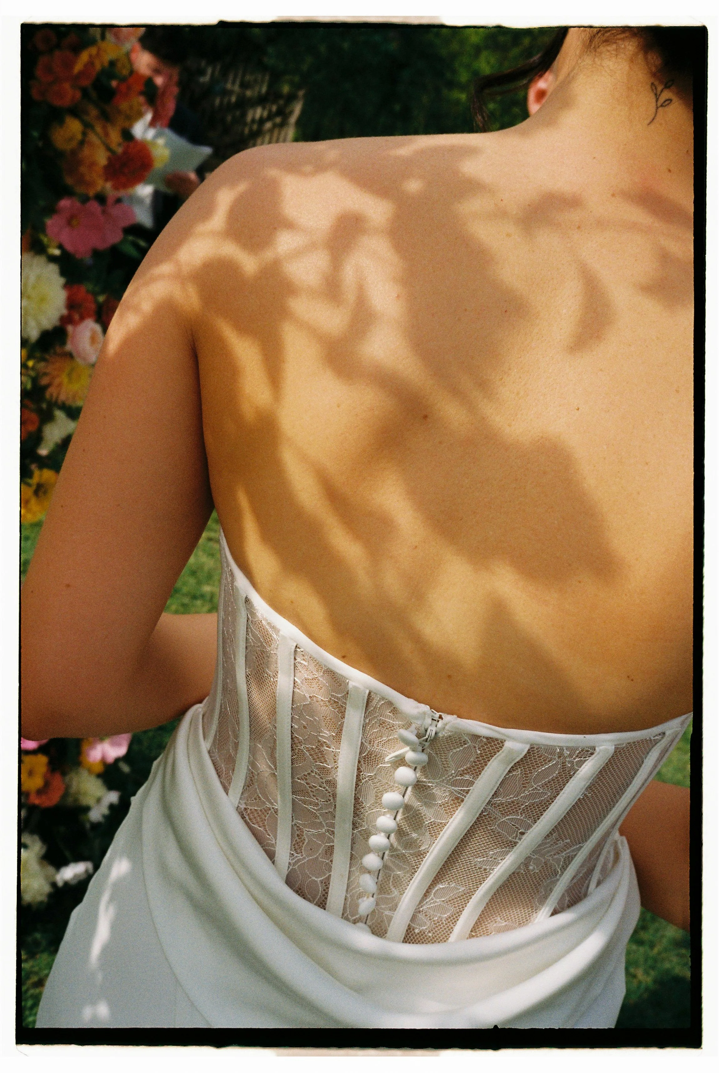 Back of a woman wearing a white dress with lace corset, shadows of leaves on her skin, outdoors with colorful flowers in the background.