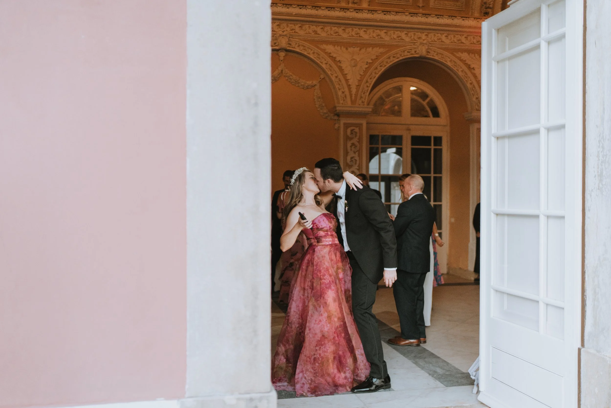 A wedding reception scene with a bride in a red gown and a groom in a black tuxedo sharing a kiss, seen through an open door. Other guests are mingling in the background inside a grand, ornate hall.
