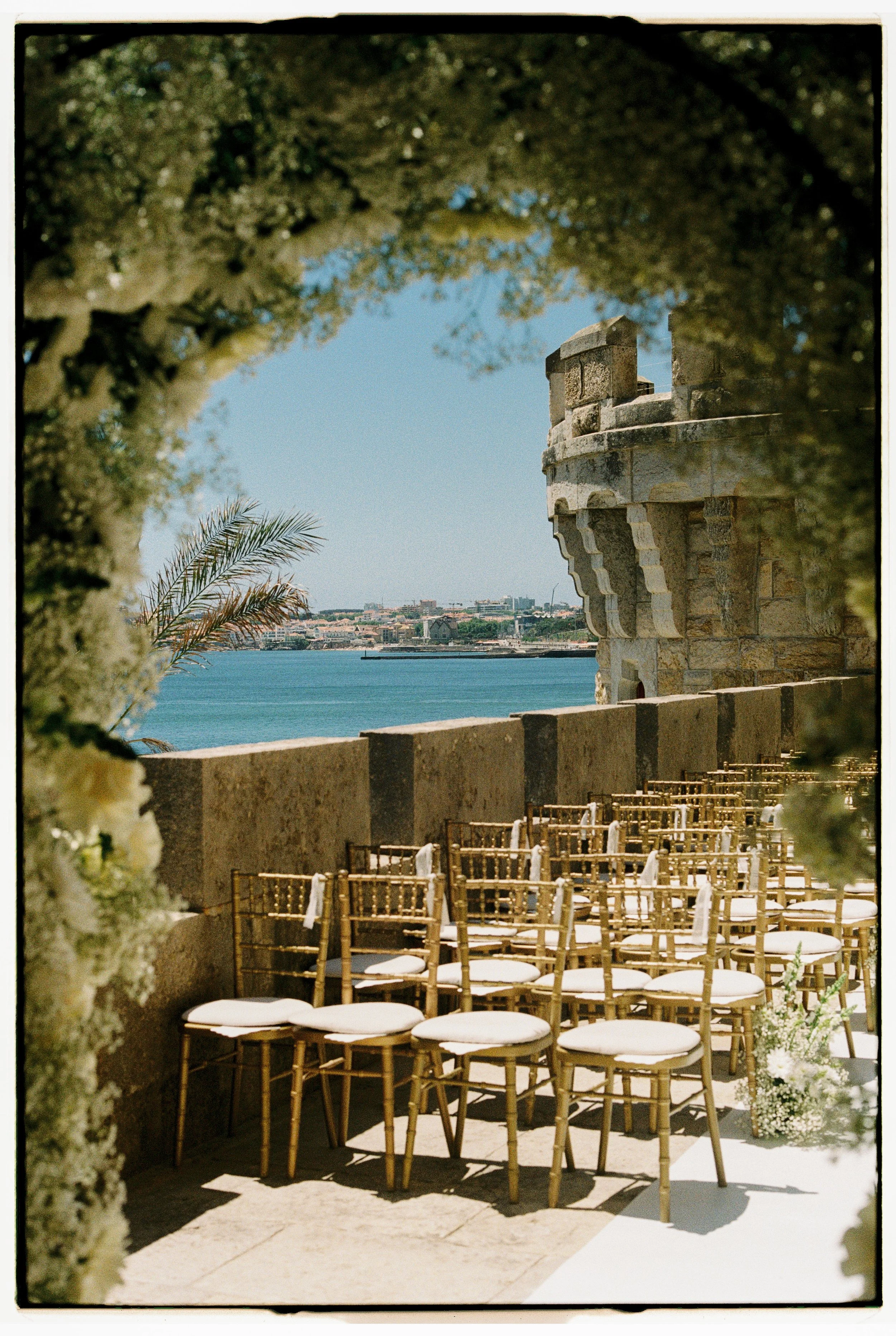 Outdoor wedding ceremony setup with chairs facing a scenic waterfront view, decorated with flowers and framed by greenery and historic stone architecture.