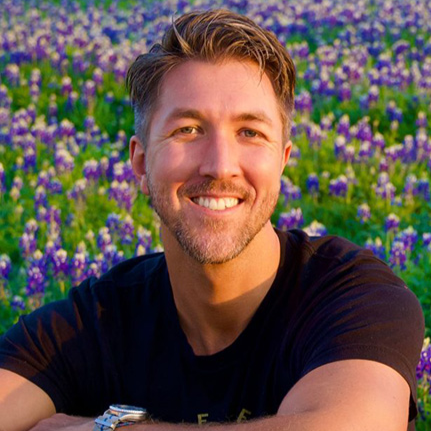 A smiling man with short brown hair and a beard, wearing a black t-shirt, standing in a field of purple and green flowers.