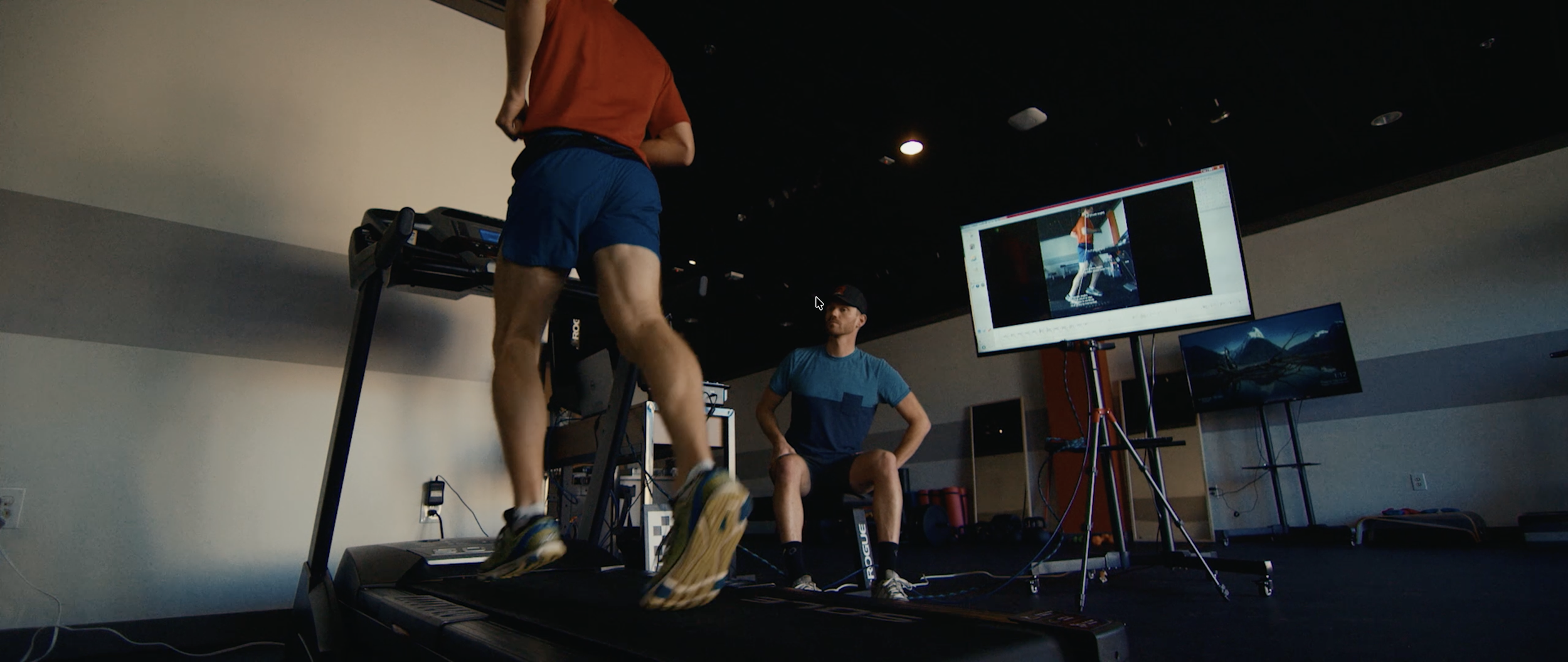 A person running on a treadmill in a lab setting, monitored by a researcher sitting nearby, with large screens displaying images and data related to the running activity.