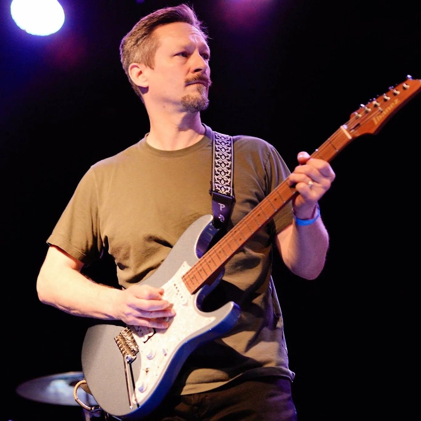 A man playing an electric guitar on stage with purple and white lighting.