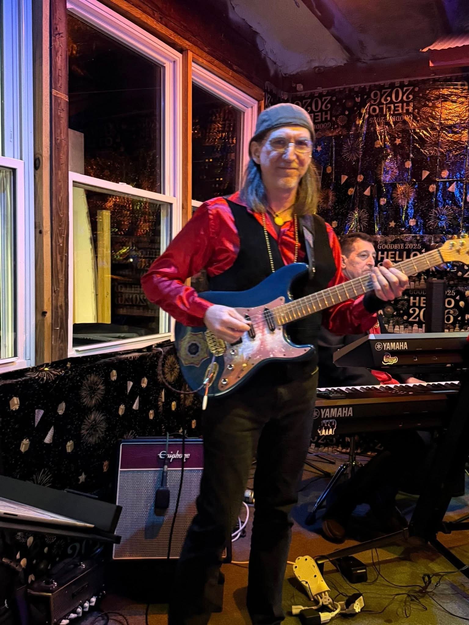 A woman with glasses, wearing a gray bandana, red shirt, and black vest, playing an electric guitar during a live music performance in a cozy venue decorated with fireworks-themed posters and black curtains.