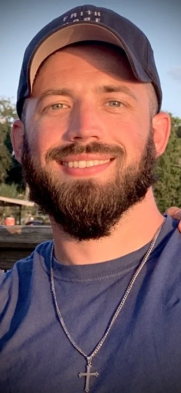 Close-up of smiling man with a beard, wearing a blue baseball cap and a chain with a cross pendant outdoors.