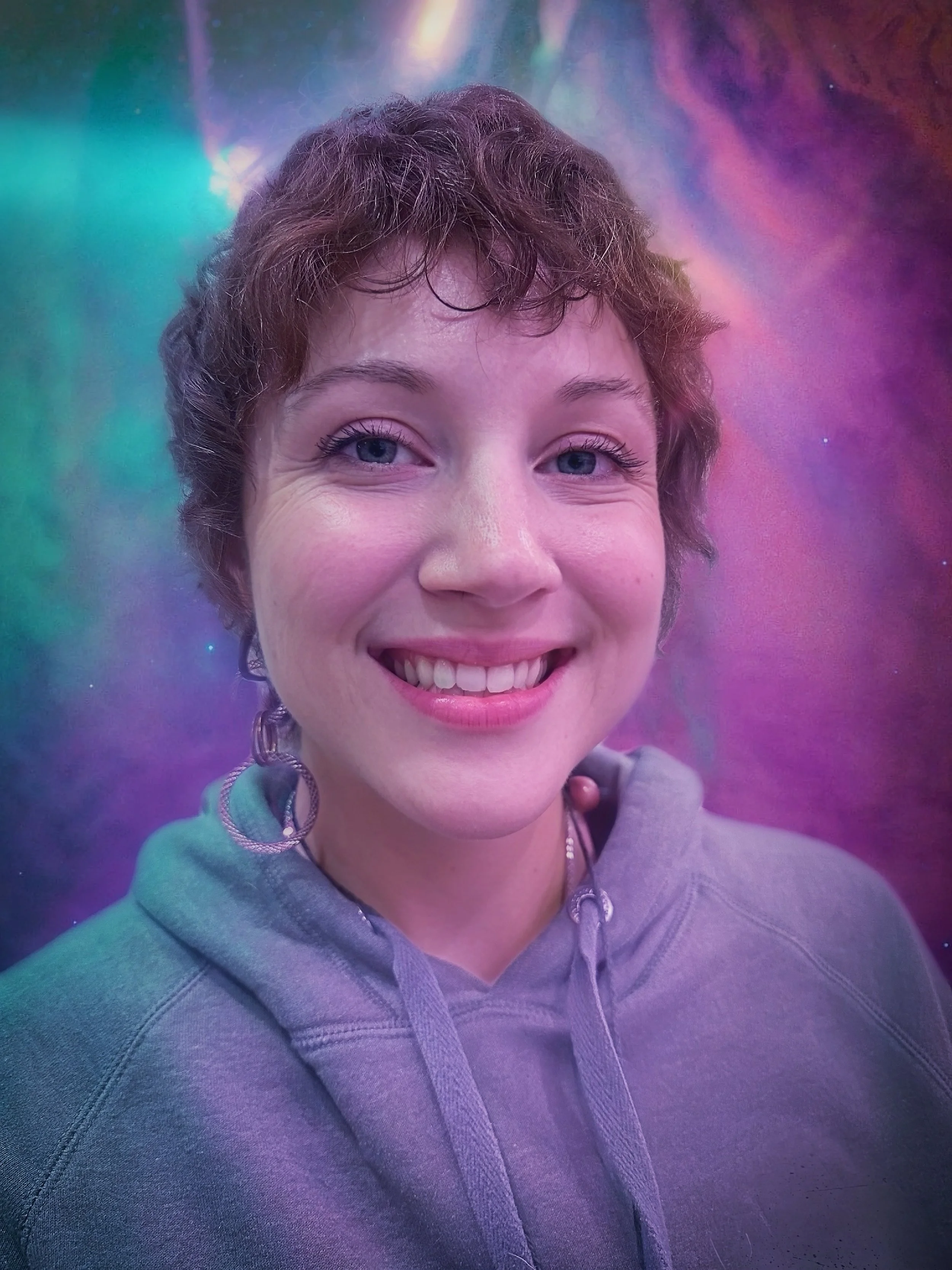 Close-up of a smiling woman with short curly hair, wearing hoop earrings and a gray hoodie, against a colorful, abstract background.
