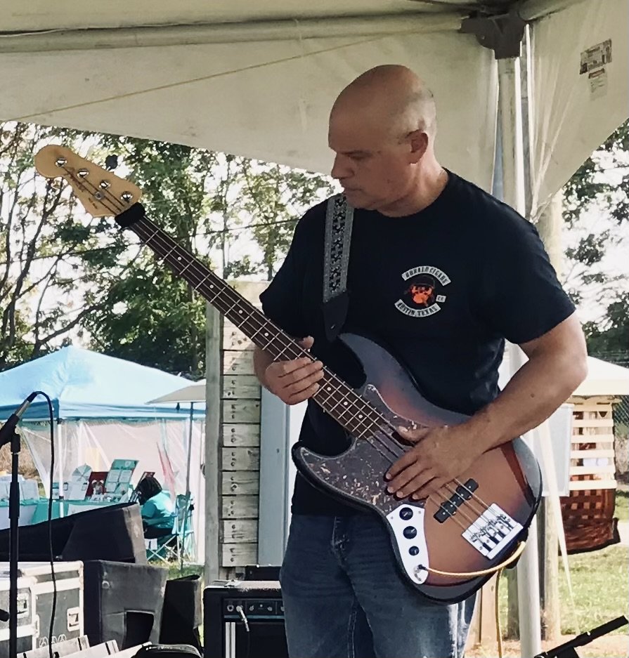 A bald man in a black t-shirt playing an electric bass guitar on an outdoor stage under a tent.