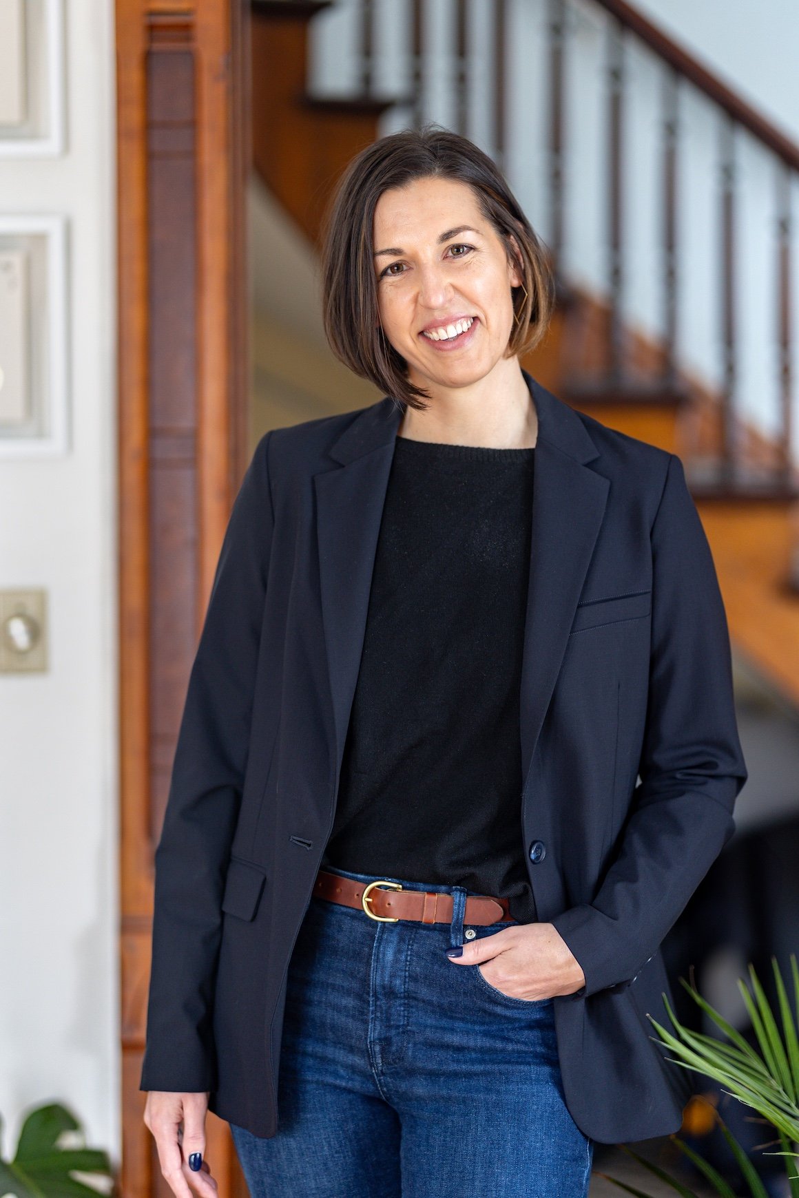 Client, Kat Nopper, owner of the interior design business, Harlan & Hazel. She has shoulder-length brown hair, wearing a navy blazer, black top, and blue jeans, standing indoors in front of a staircase with a wooden railing.