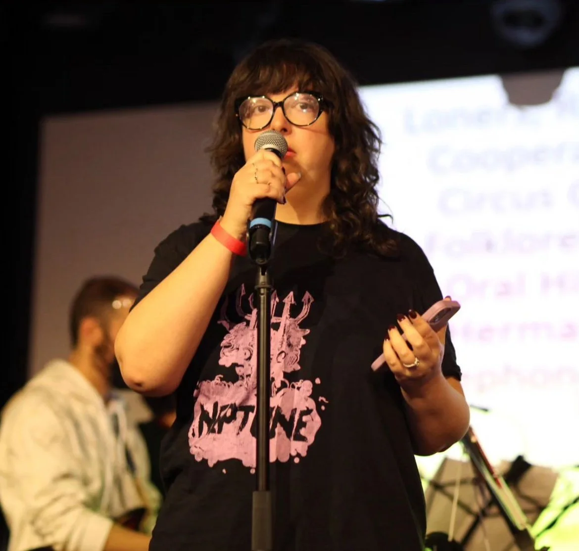 Client, Keira Horowitz, a Boston-based writer and speaker at an event, speaking into a microphone on a stage with a presentation behind her.