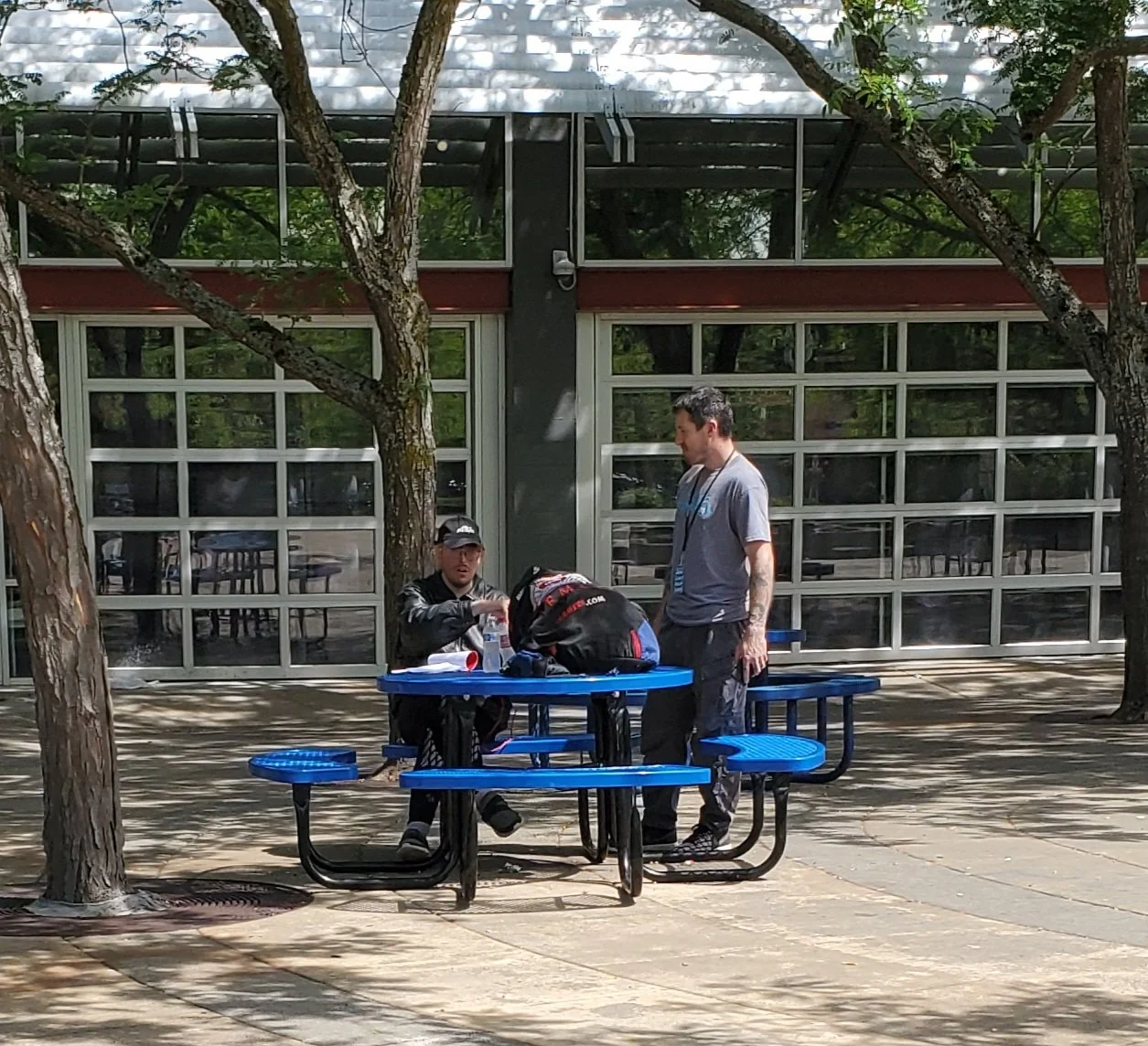 Two men are having a conversation at a blue picnic table outside a building with large glass windows. One man is seated, wearing a black jacket and baseball cap, while the other man stands, wearing gray clothing with tattoos on his arms. Trees provid