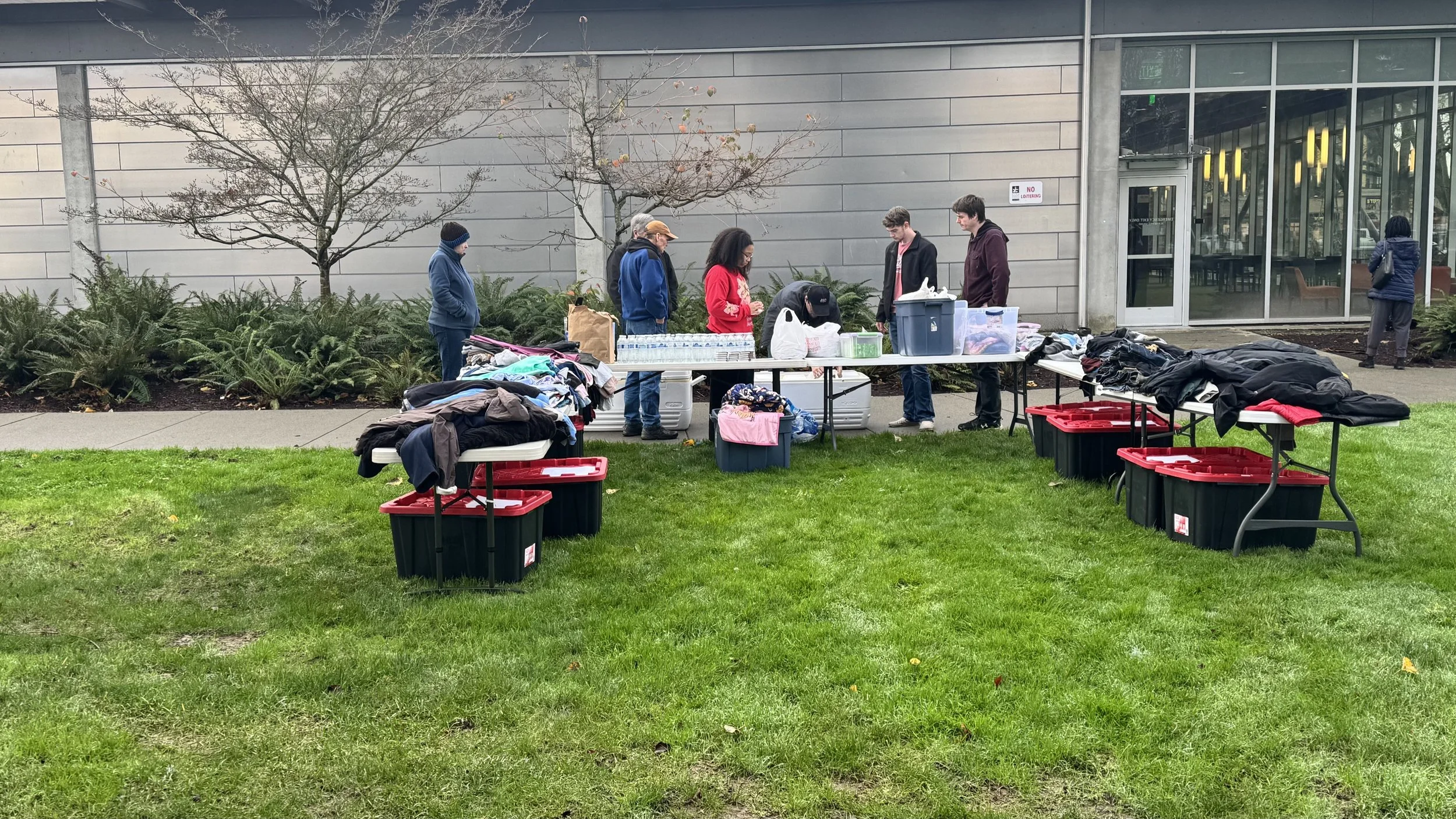 People shopping at an outdoor clothing sale with tables of folded clothes in front of a modern building.