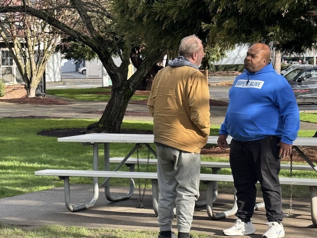 Two men talking in a park. One man is wearing a yellow jacket, gray pants, and black shoes. The other is wearing a blue hoodie with white text, black pants, and white shoes. They stand near a white picnic table under a large tree.