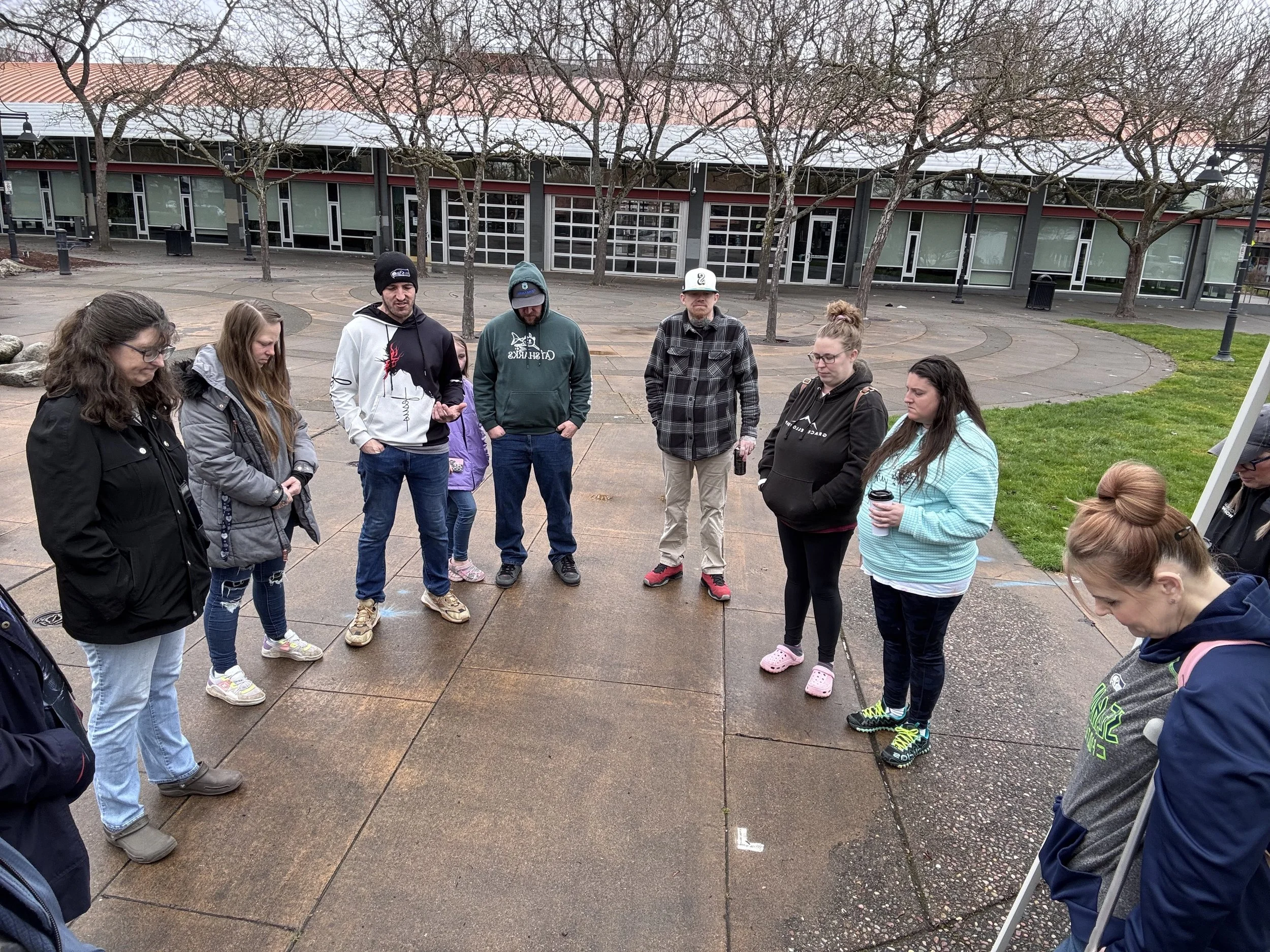 A group of people standing in a circle outdoors on a wet pavement, some with heads bowed, possibly in prayer or reflection, with a building and trees in the background.