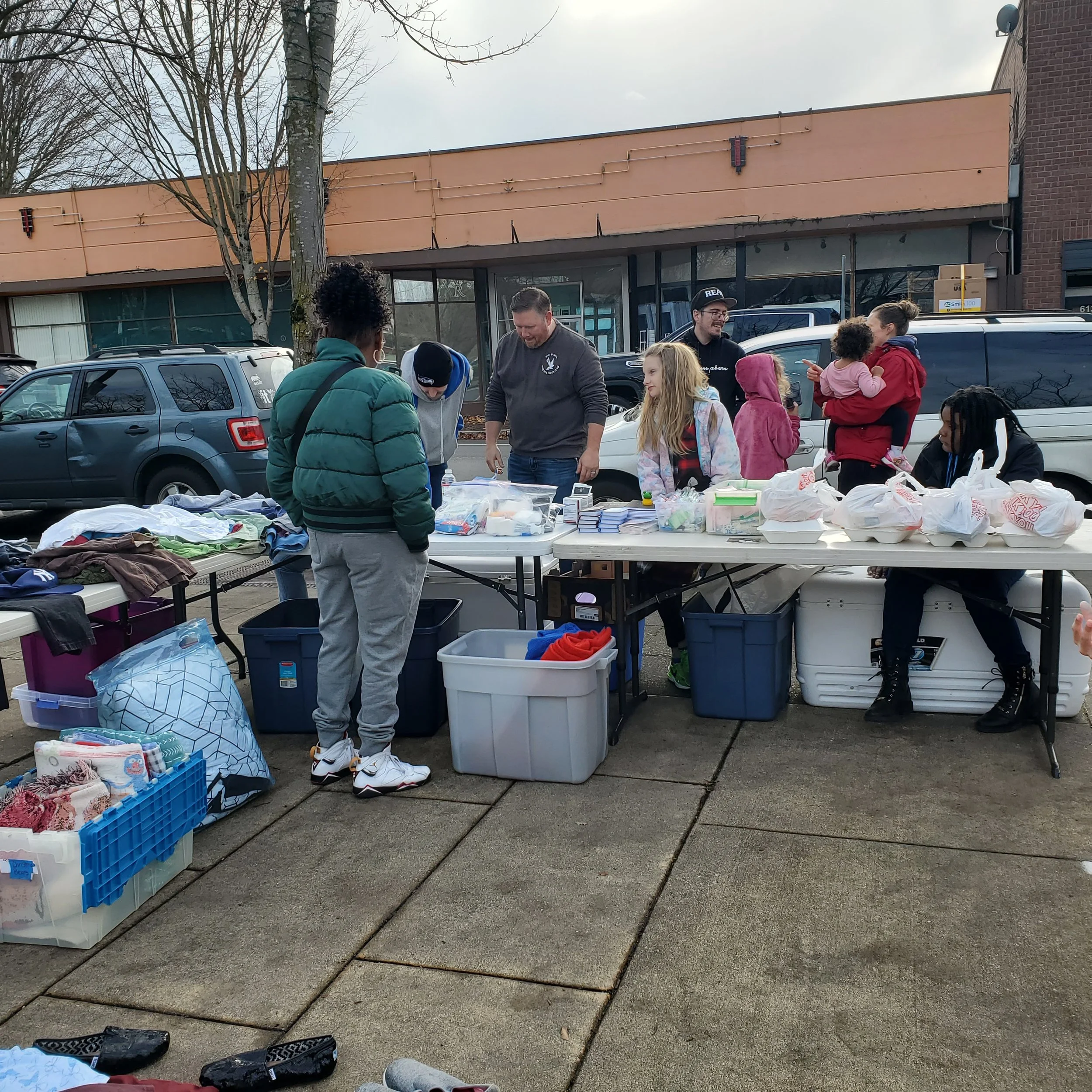 People at an outdoor table selling clothing and other items in a parking lot. Several vehicles are parked behind the table, and a building is in the background. Some items are in bins on the ground.