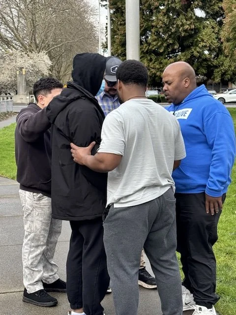 Group of five men standing closely together outdoors, praying together.
