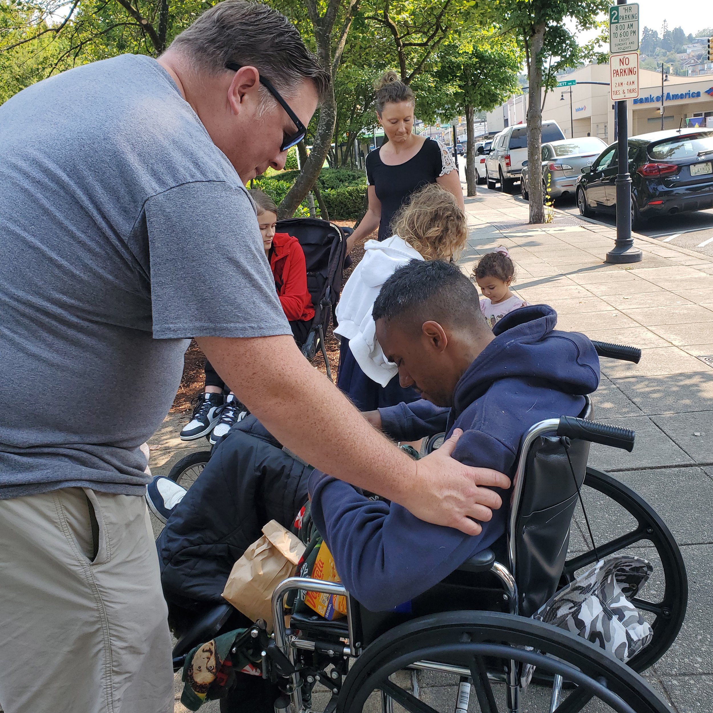 A man leaning over to comfort a young man in a wheelchair on a sidewalk, with several other people including women and children in the background, and parked cars along the street under trees.