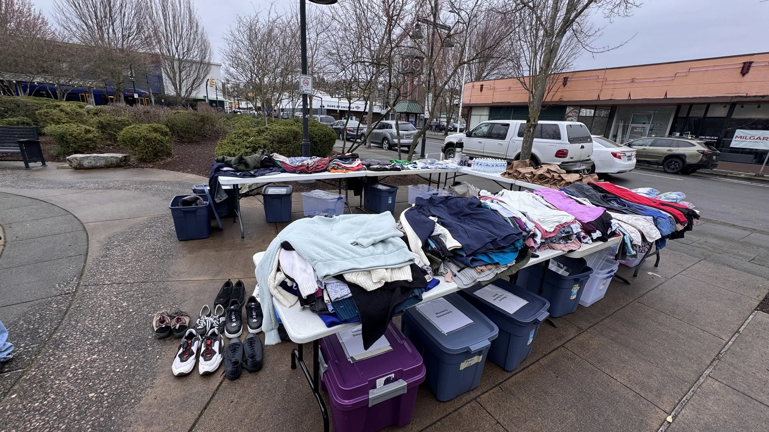 Tables set up on a sidewalk displaying clothing, shoes, water bottles, and miscellaneous items for sale during an outdoor market or yard sale on a cloudy day.