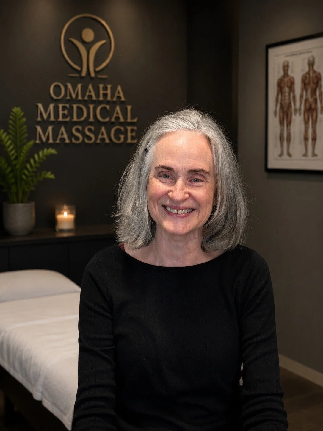 A smiling woman with gray hair sitting in a massage therapy room at Omaha Medical Massage, with a treatment bed, a candle, a potted plant, and framed anatomical posters on the wall.