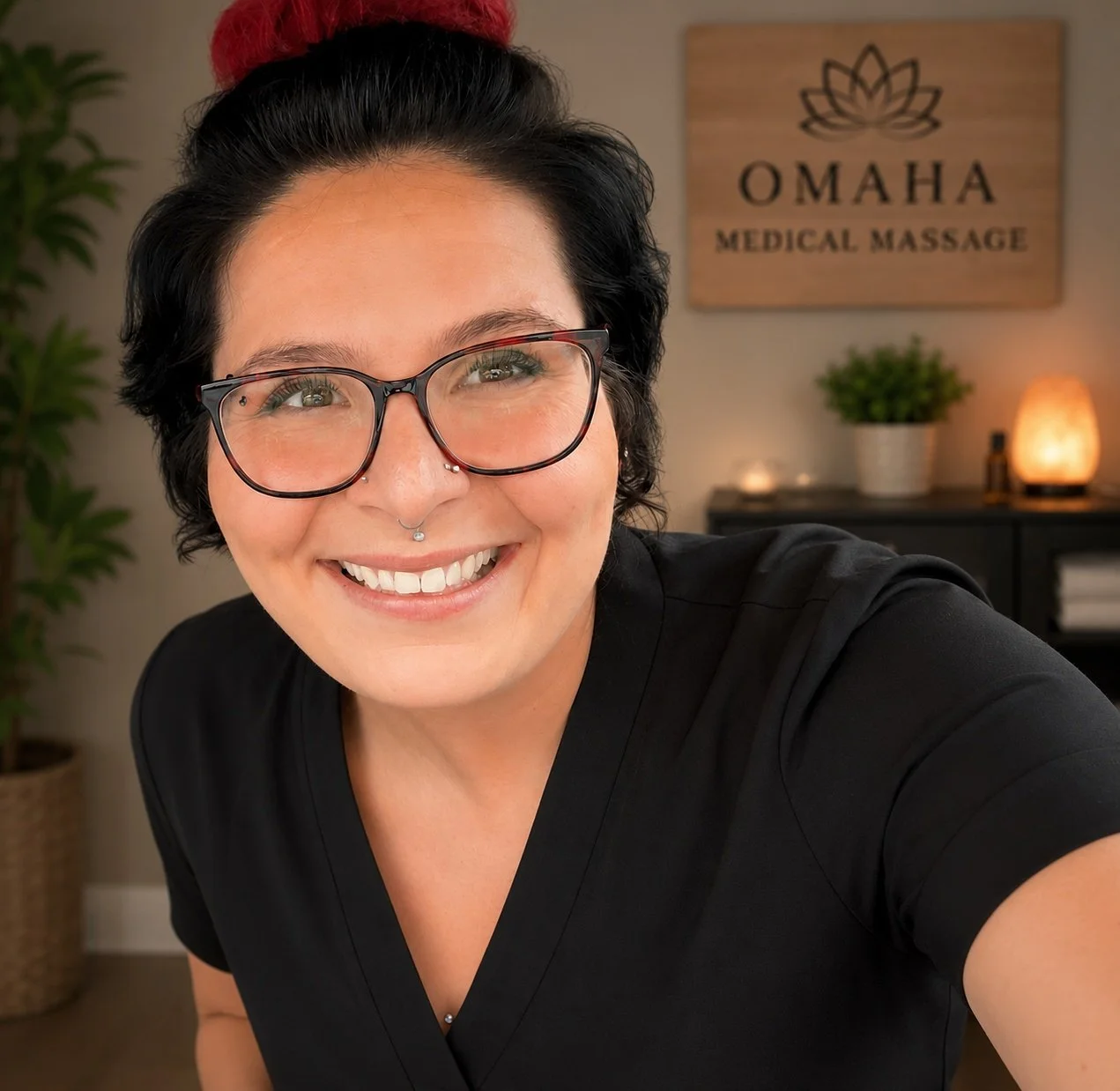 A woman with short black hair and glasses smiling for a selfie in a wellness or massage clinic named Omaha Medical Massage, with plants and dim lighting in the background.