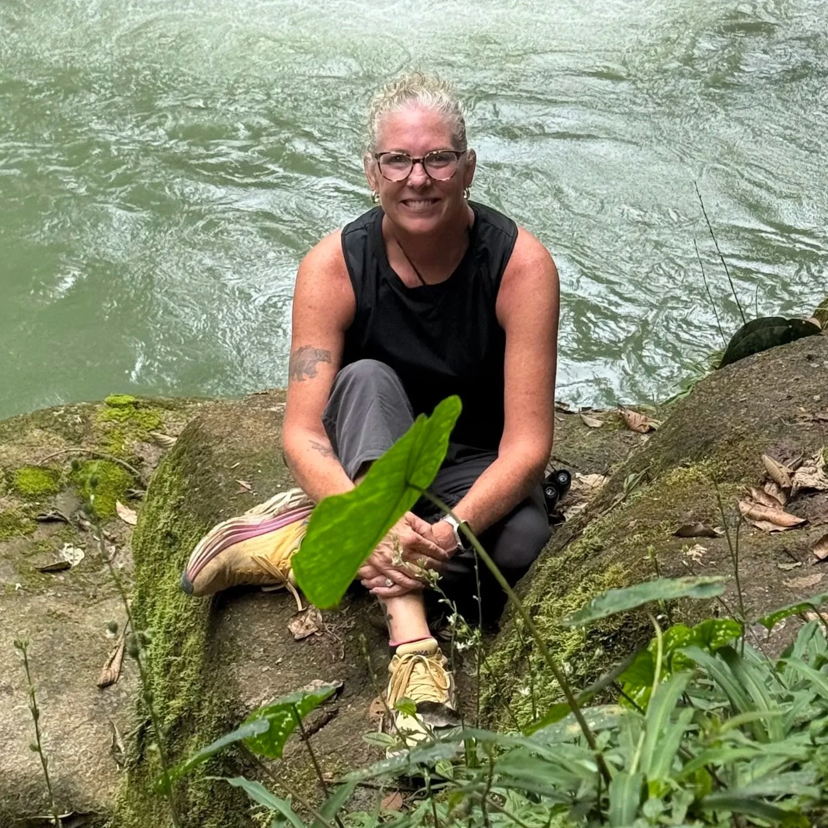 A woman sitting on a moss-covered rock in a riverside setting, smiling at the camera, wearing glasses, a black sleeveless shirt, gray pants, and yellow shoes, with greenery in the foreground and water in the background.